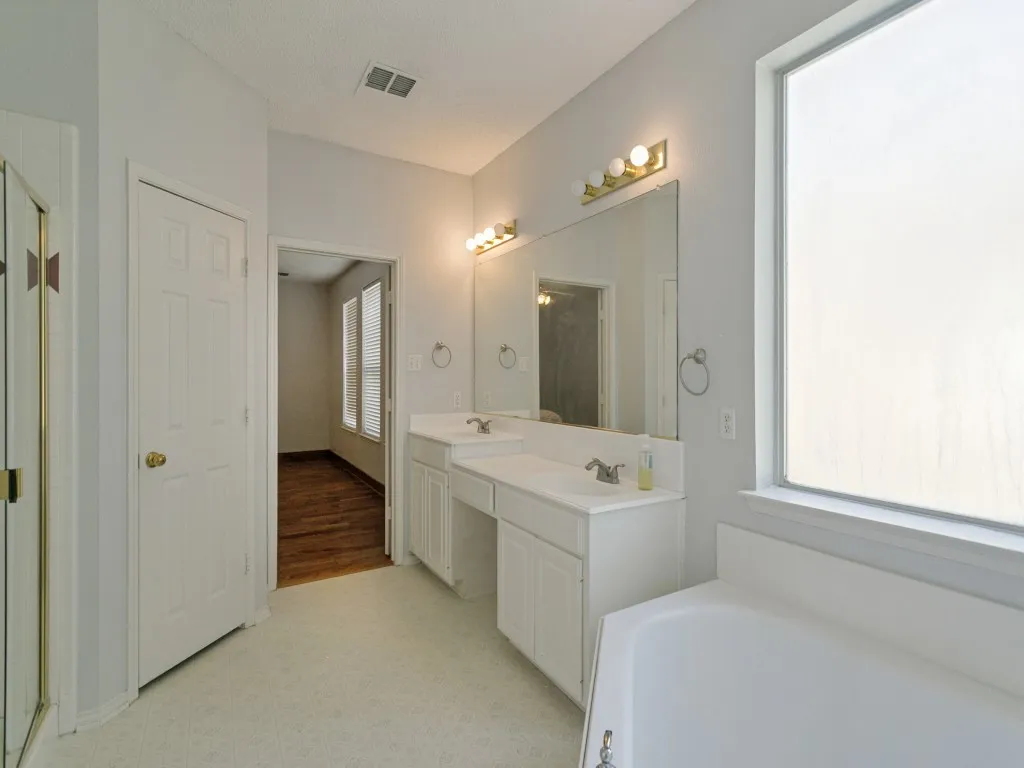 Dual vanity with white cabinetry and integrated sinks, large mirror with vanity lighting, and an oversized soaking tub
