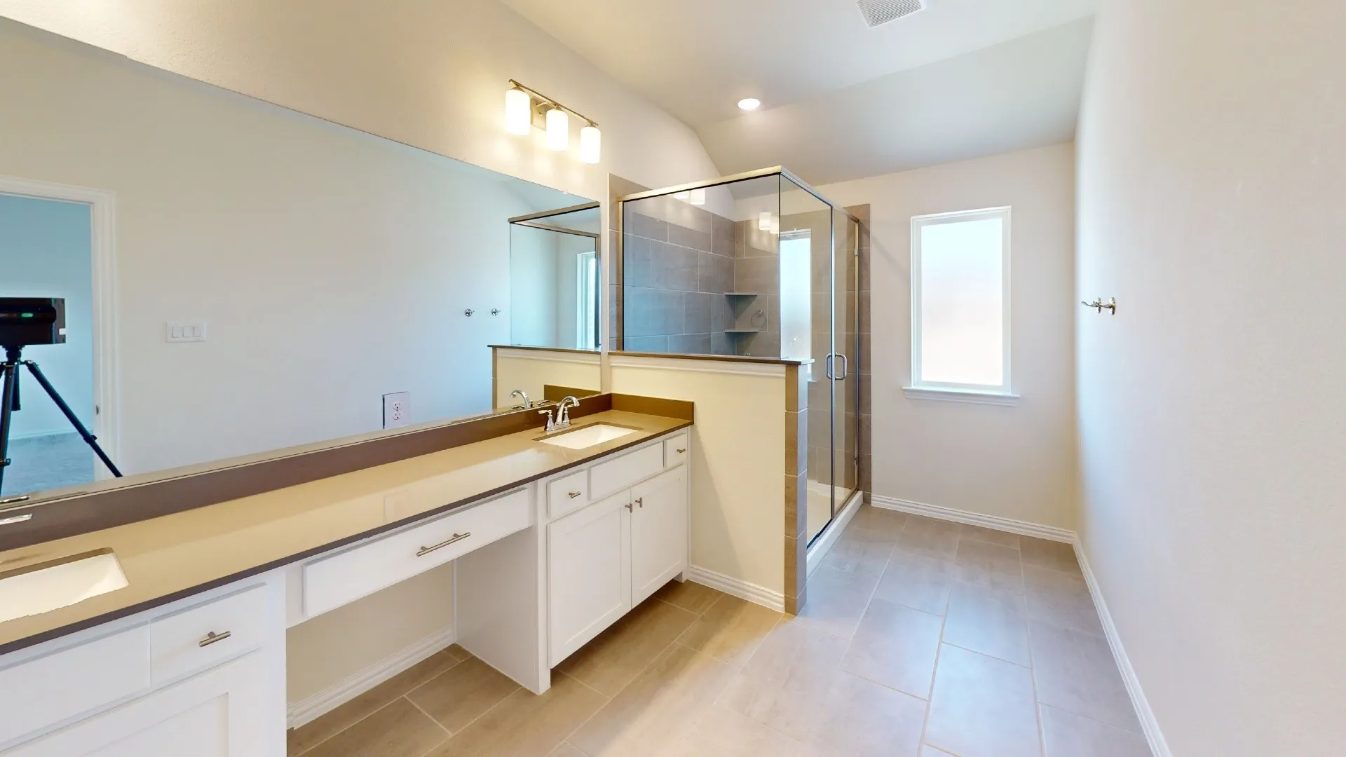 Dual vanity with integrated sinks, expansive mirror, full-size shower with glass enclosure, tiled shower surround with built-in shelving, and neutral floor tile