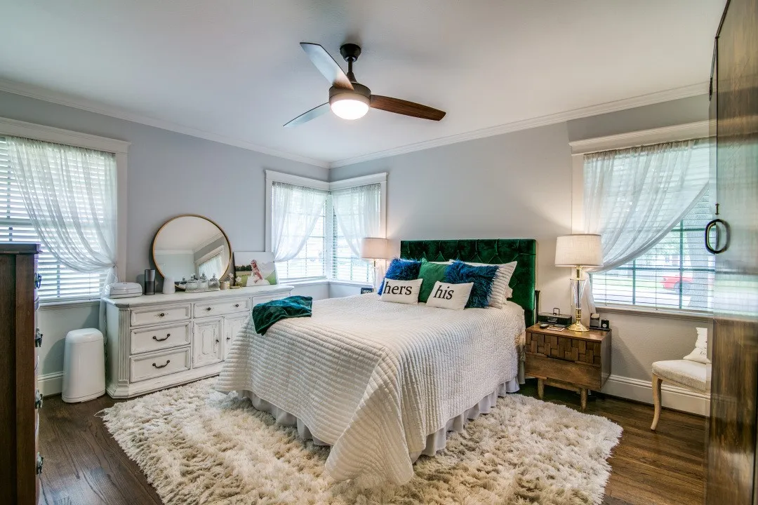 Wood-finish flooring, corner windows with blinds, ceiling fan with integrated lighting, built-in dresser, and a velvet upholstered headboard