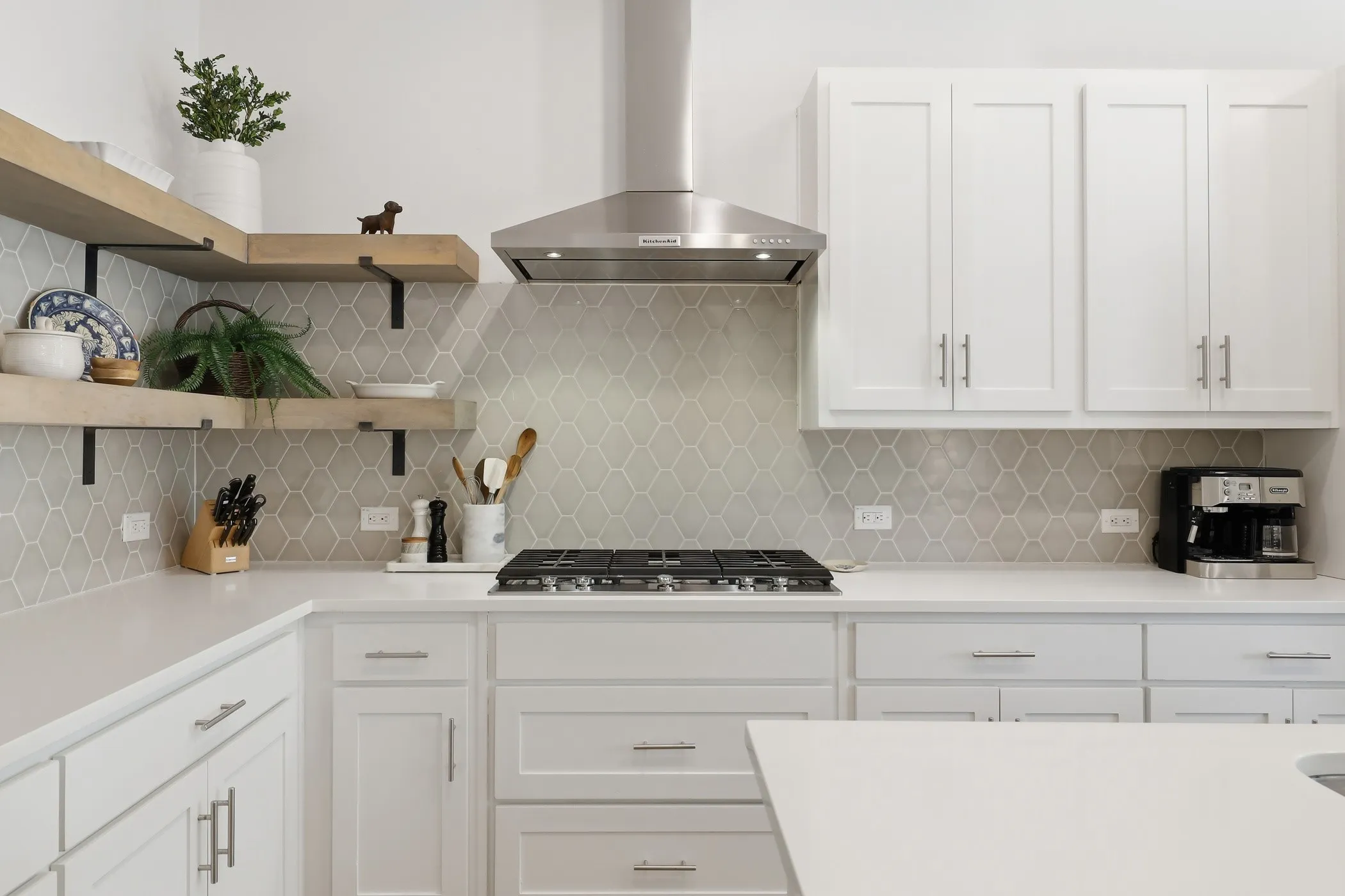 White cabinetry with brushed nickel hardware, light-toned countertops, geometric pattern backsplash, stainless steel range hood, and open wood-finish shelving