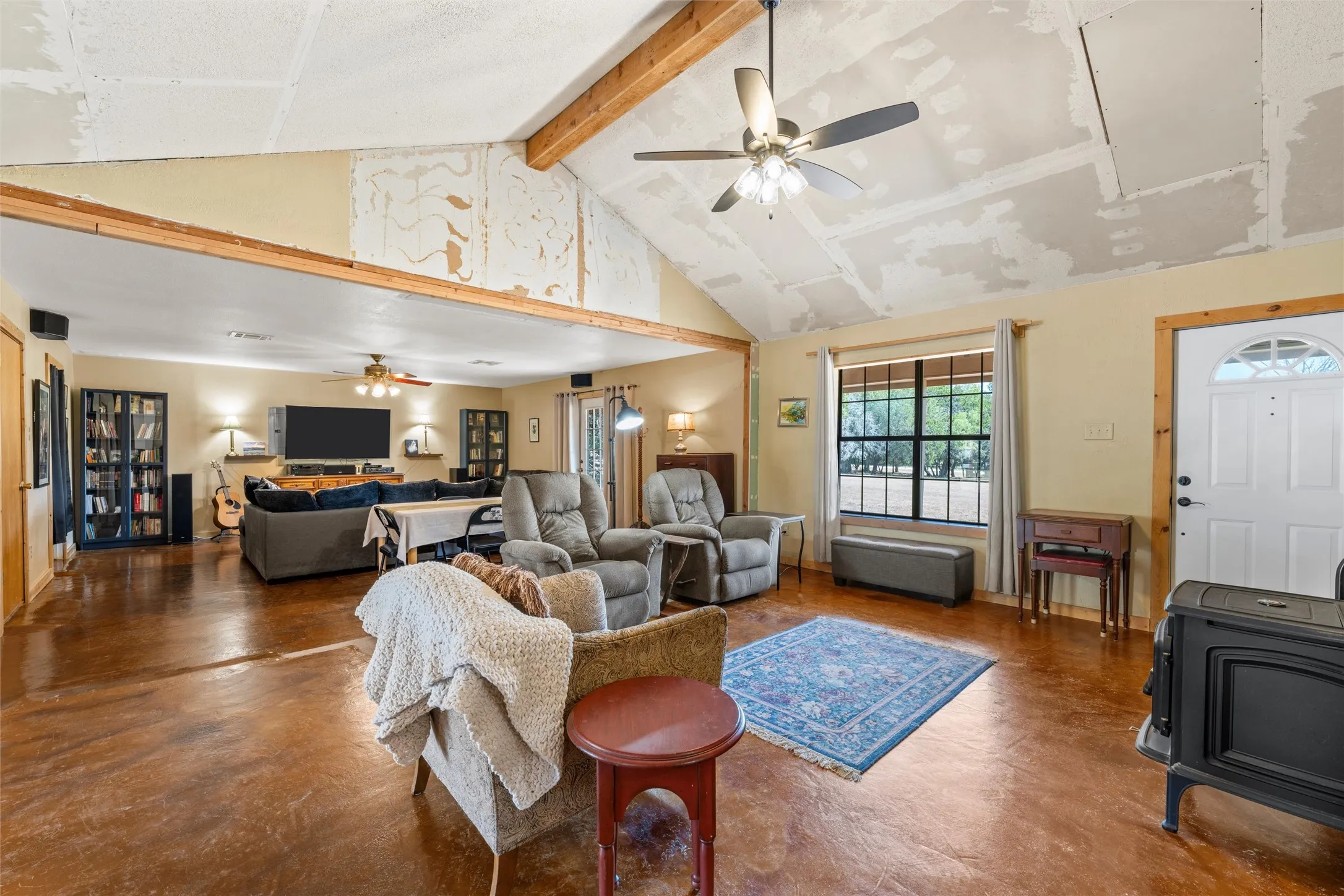 Vaulted ceiling with exposed wood beams, stained concrete flooring, recessed wood-framed window, entry door with arched lites, and a freestanding wood-burning stove