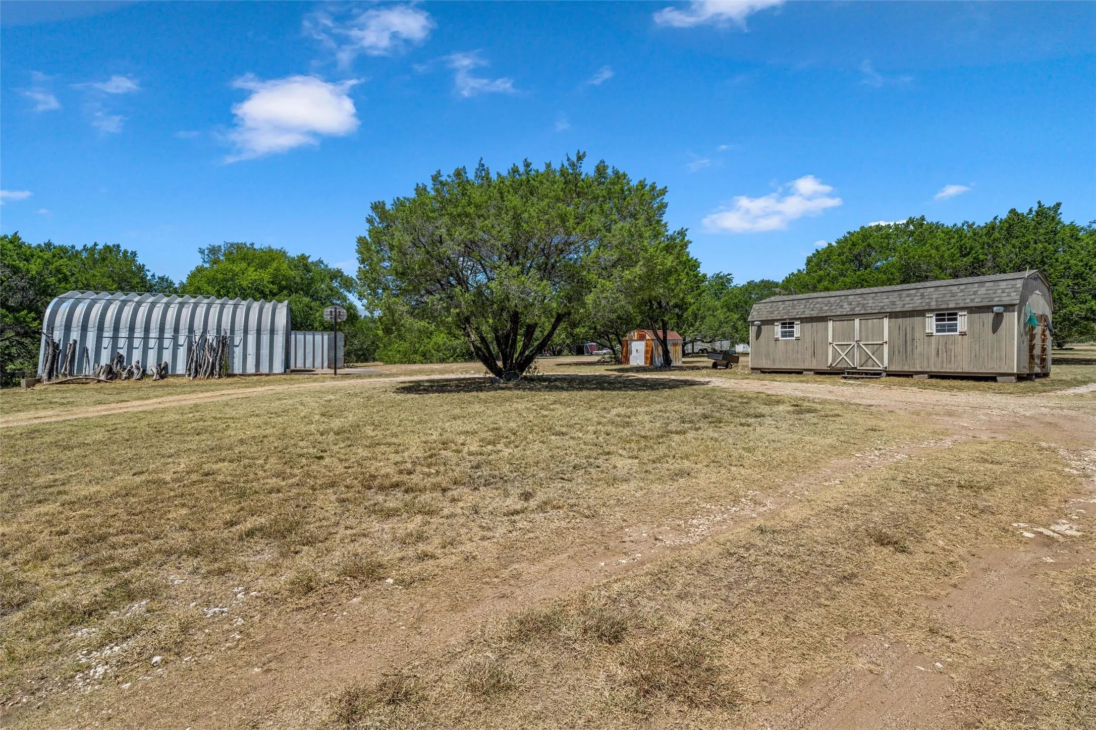 Metal Quonset hut structure, detached shed with shingle roof, mature trees, and a gravel surface