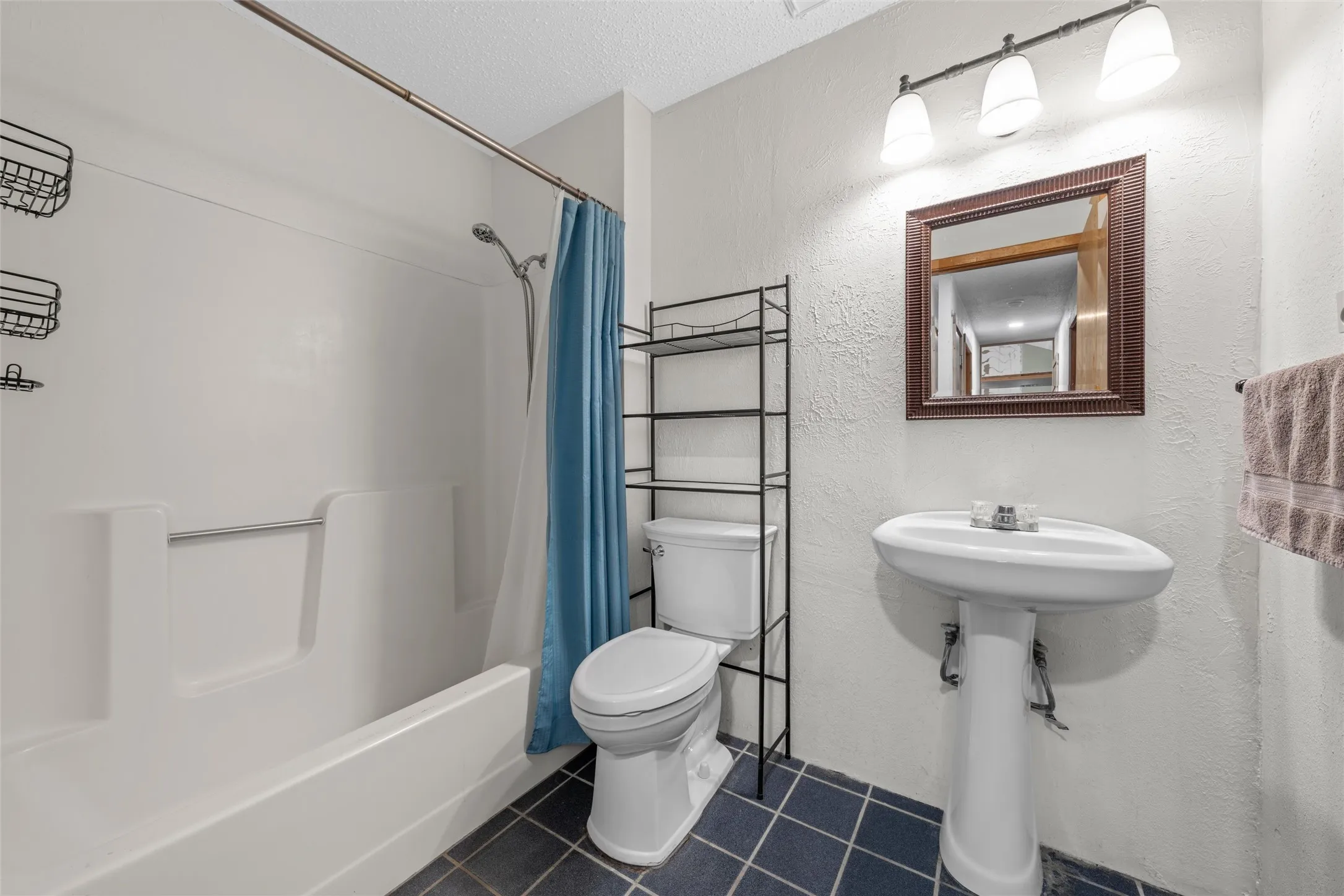 White tub and shower combination, pedestal sink, wall-mounted mirror, three-light vanity fixture, and tiled flooring