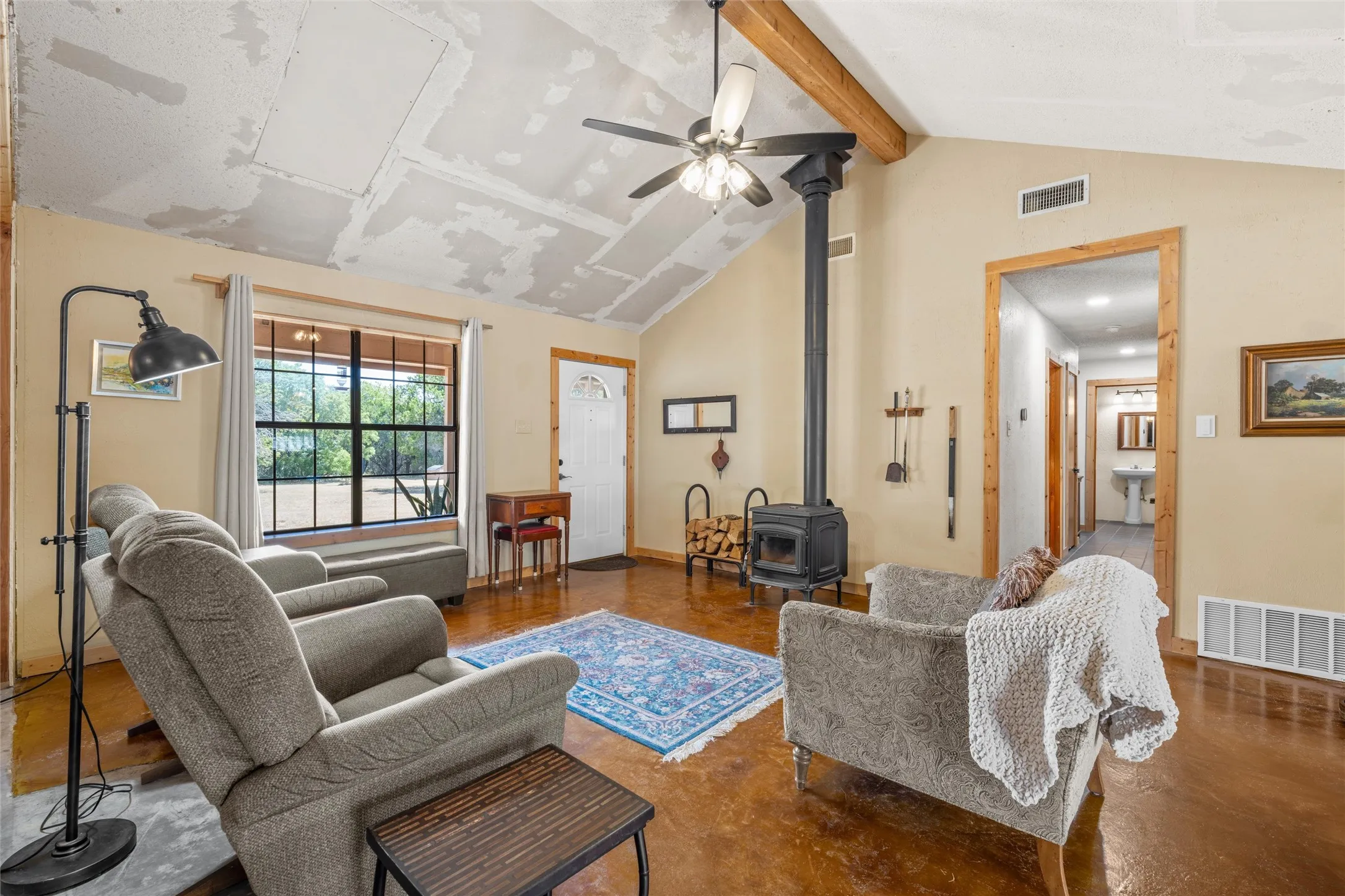 Vaulted ceiling, exposed wood beam, wood-burning stove, wood-finish flooring, and a large multi-pane window