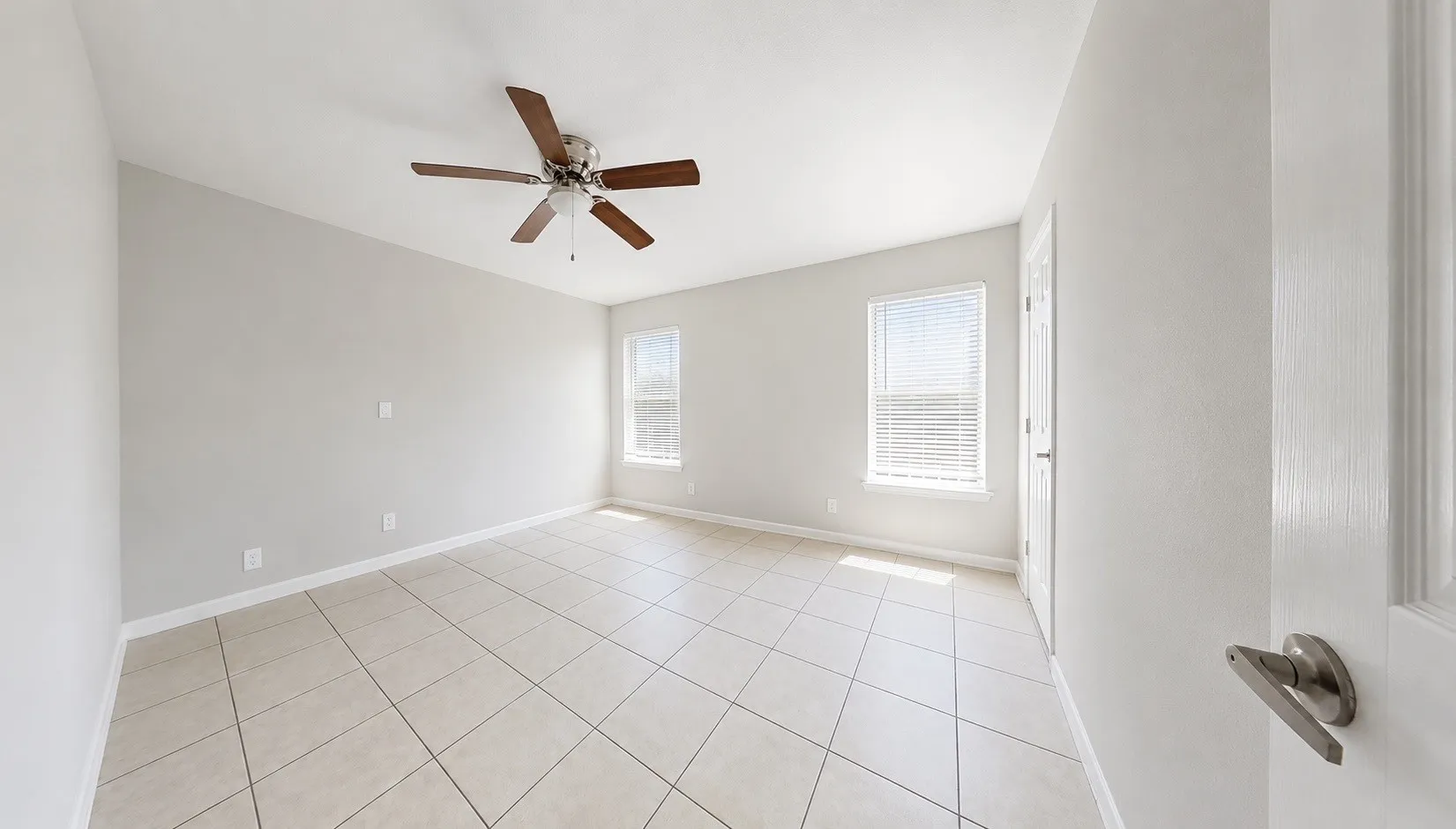 Spacious room featuring neutral tone walls, tile flooring, two windows with blinds, a ceiling fan, and a paneled door with brushed nickel hardware