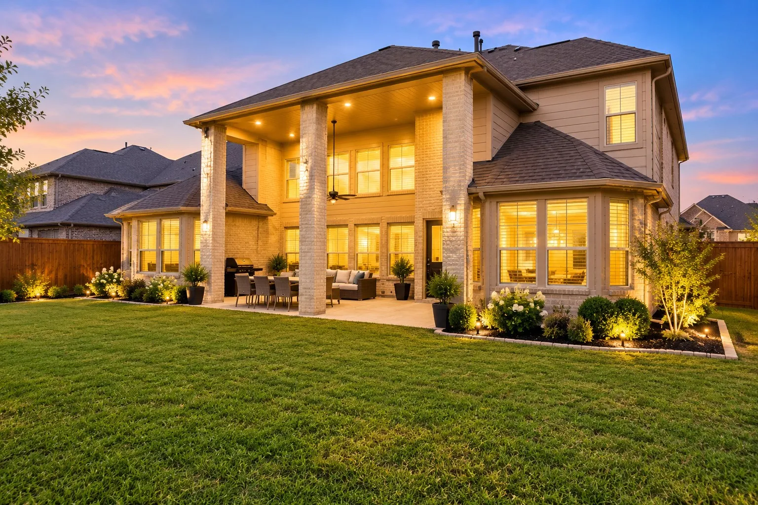 Expansive backyard featuring a covered patio, brick columns, and a manicured lawn with strategic landscaping