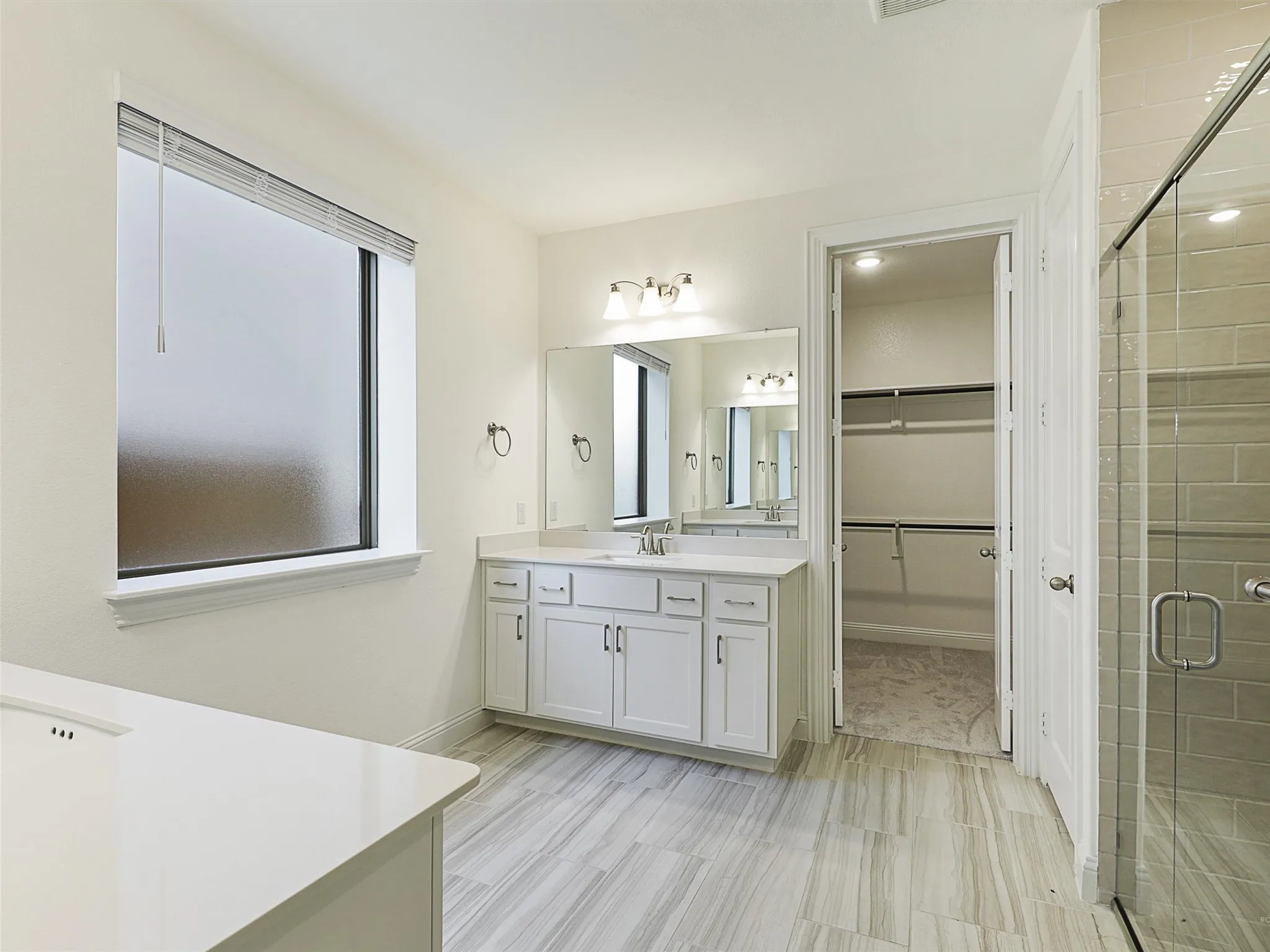 Spacious bathroom featuring a double vanity with ample storage, a glass-enclosed shower, wood-finish tile flooring, and a walk-in closet with built-in shelving
