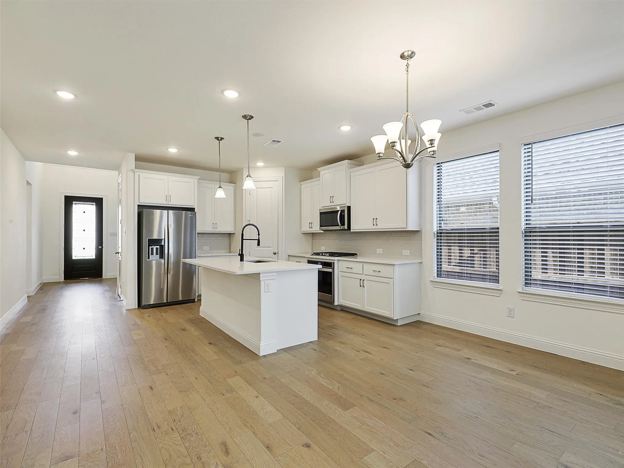 Open-concept living area featuring wood-finish flooring, a kitchen island with pendant lighting, white cabinetry, stainless steel appliances, and a dining area with a chandelier