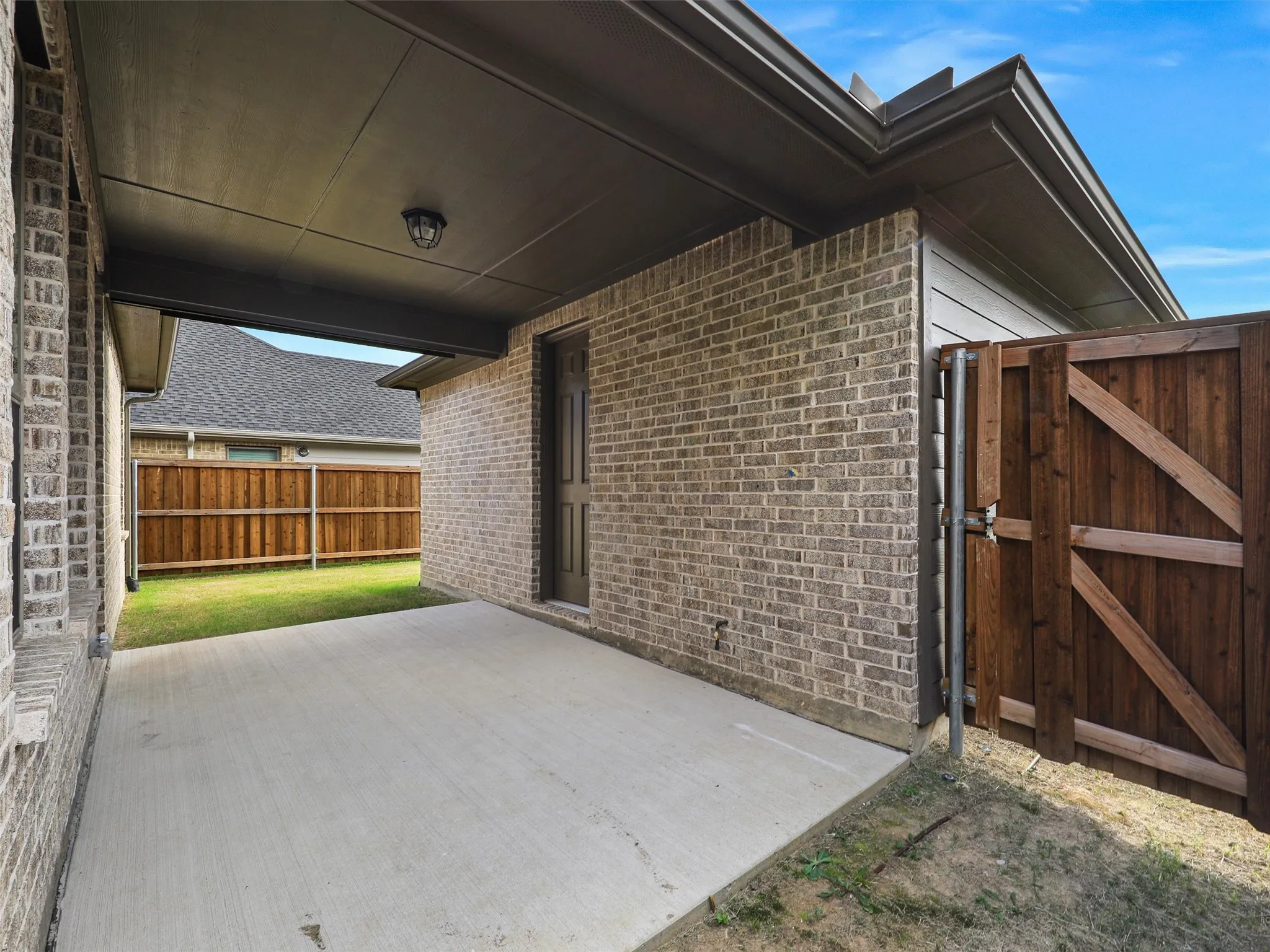 Covered concrete patio with a brick exterior, a wood privacy fence, and a dark-paneled door