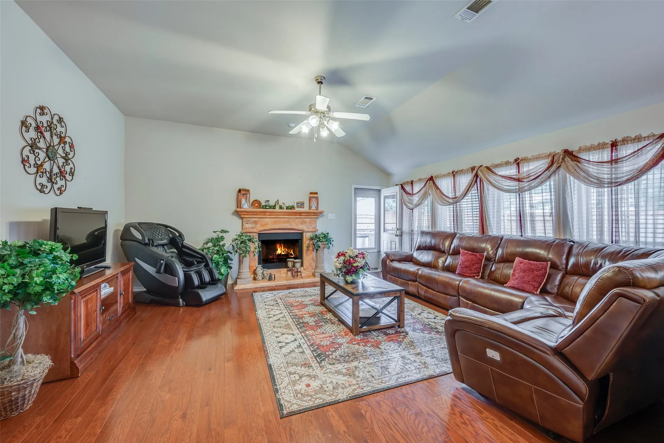 Vaulted ceiling, wood-finish flooring, fireplace with decorative mantel, ceiling fan with light fixture, and multiple large windows
