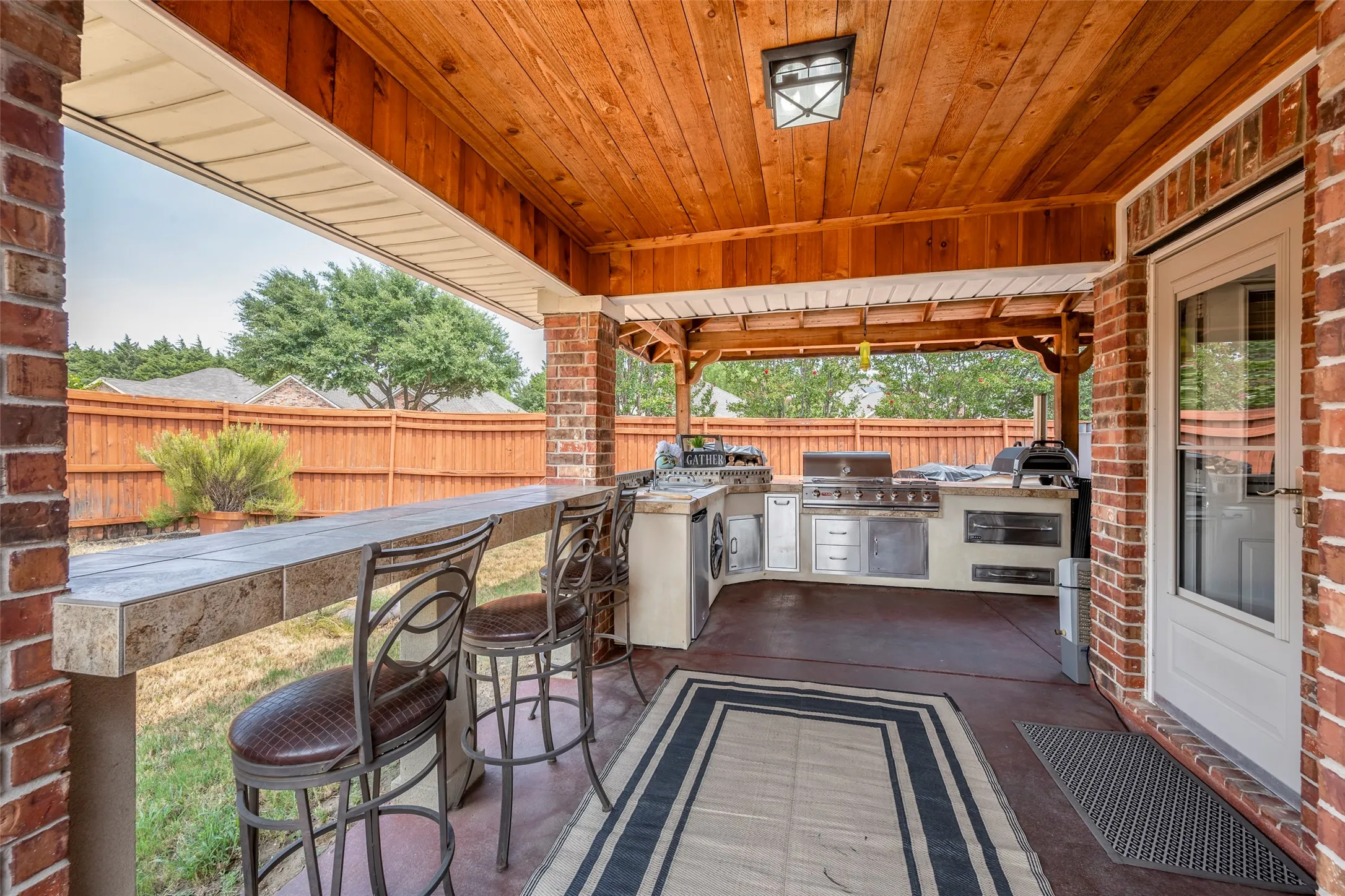 Covered patio featuring an outdoor kitchen with built-in appliances, a wood-paneled ceiling, and brick columns