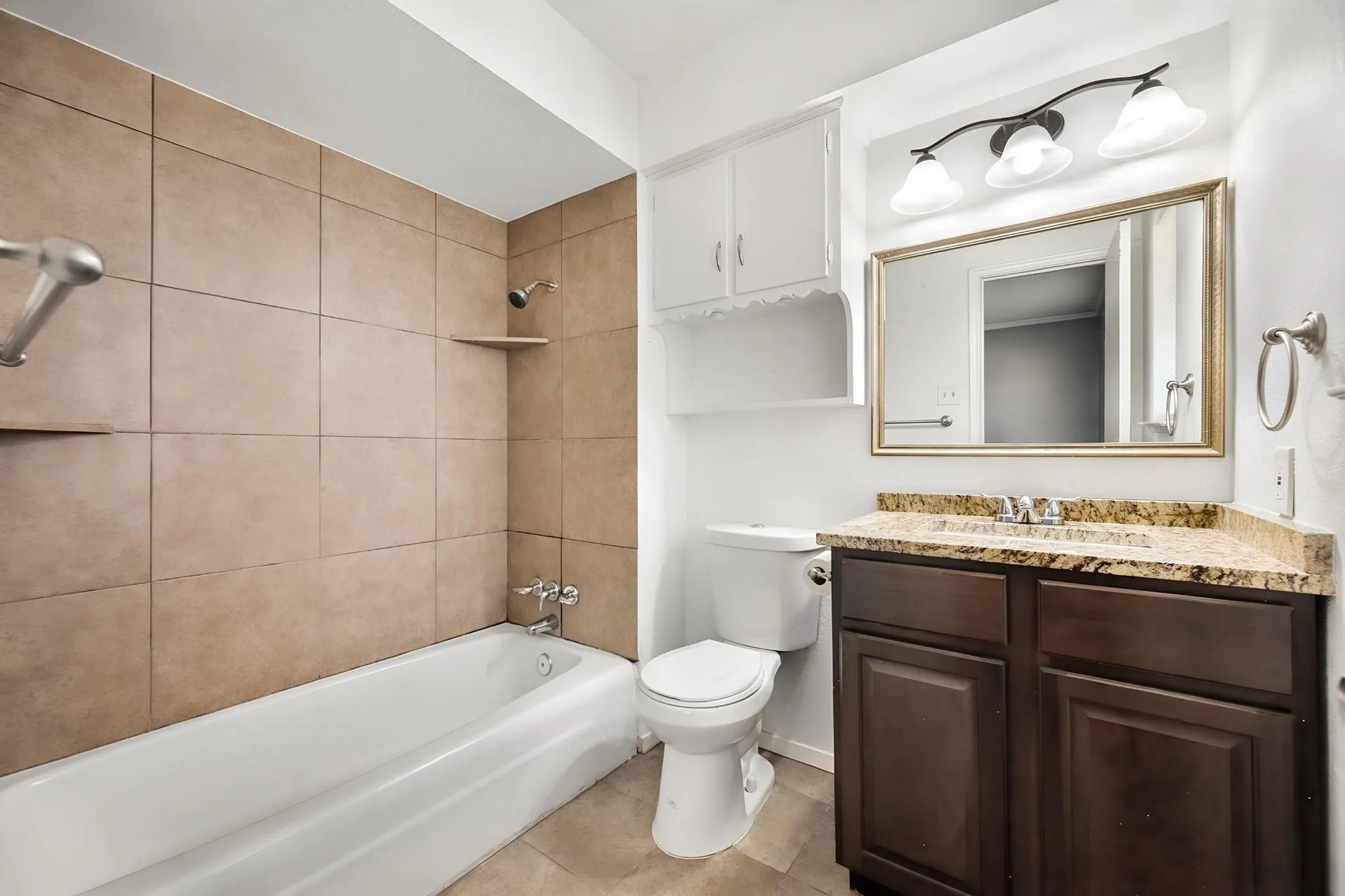 Bathtub with tile surround, built-in storage shelves, dark wood-finish vanity with stone countertop, and wall-mounted mirror