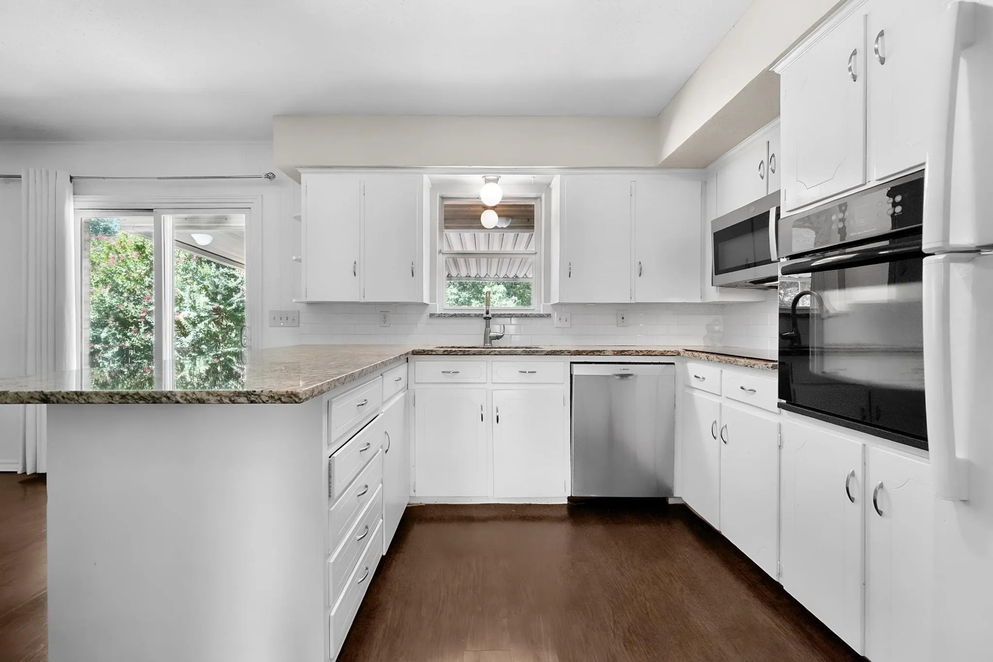 White cabinetry with silver-tone hardware, wood-finish flooring, L-shaped countertop with integrated sink, stainless steel dishwasher, and built-in stainless steel microwave and oven