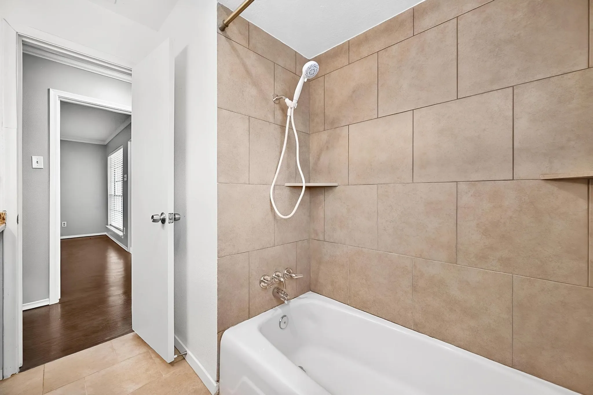 White bathtub with a chrome shower fixture, set against large format beige wall tiles and integrated shower shelves