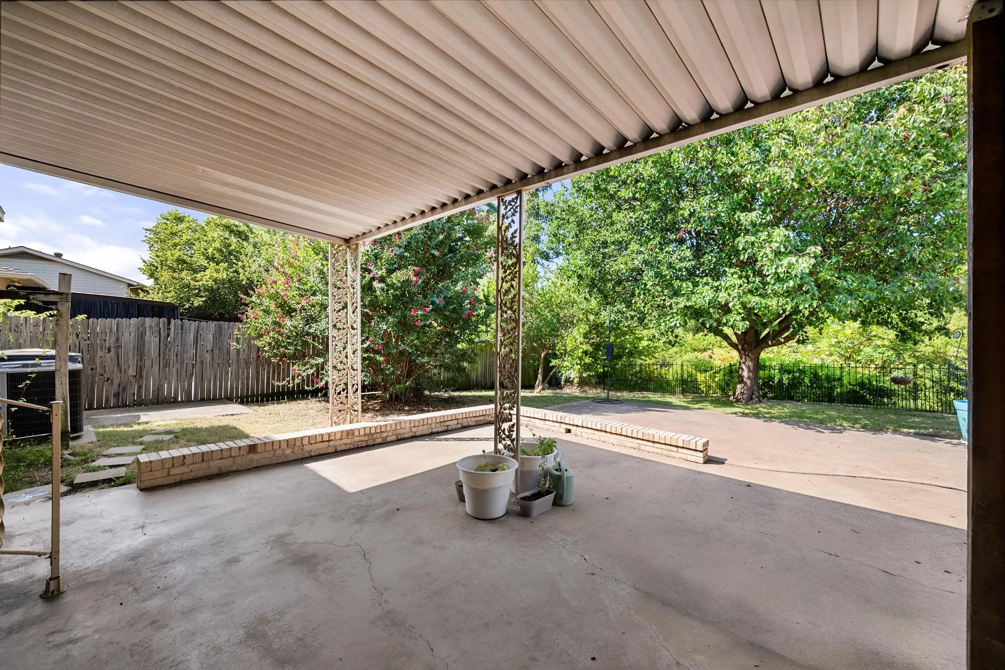 Covered patio with concrete flooring, decorative metal support columns, a brick planter, and a wood privacy fence