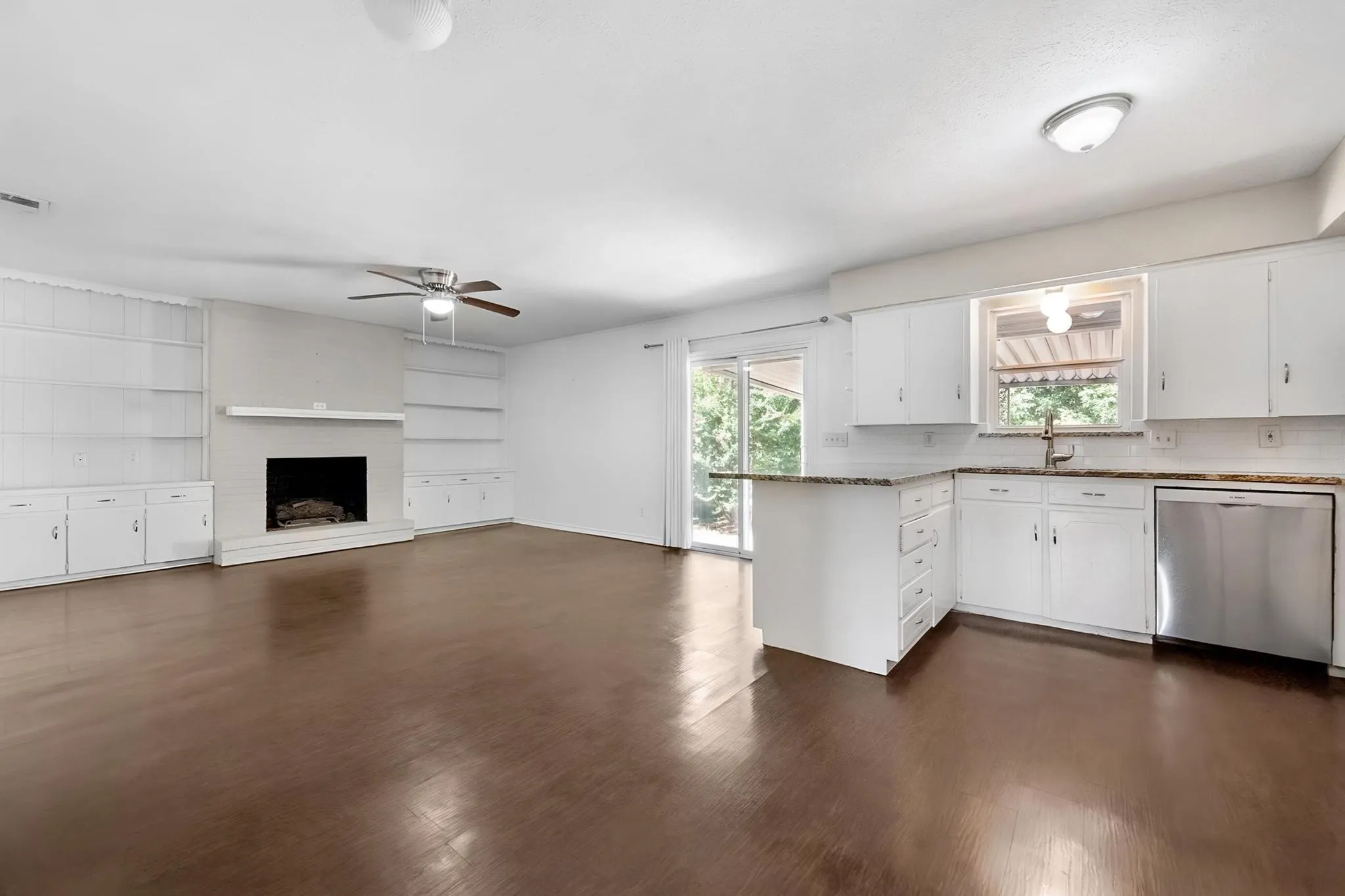 Open-concept living space with dark wood-finish flooring, a white brick fireplace, built-in shelving, white cabinetry, and stainless steel appliances