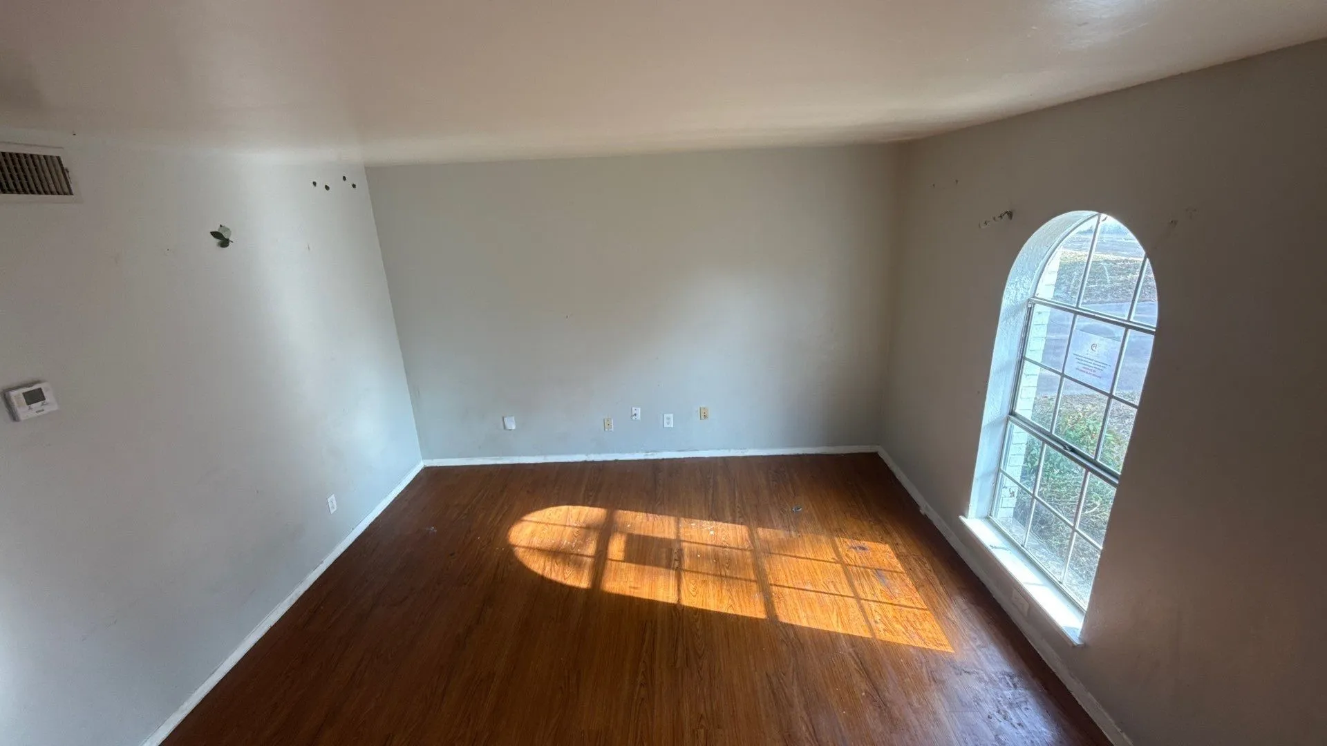 Arched window, wood-finish flooring, light gray walls, white baseboards, wall-mounted thermostat