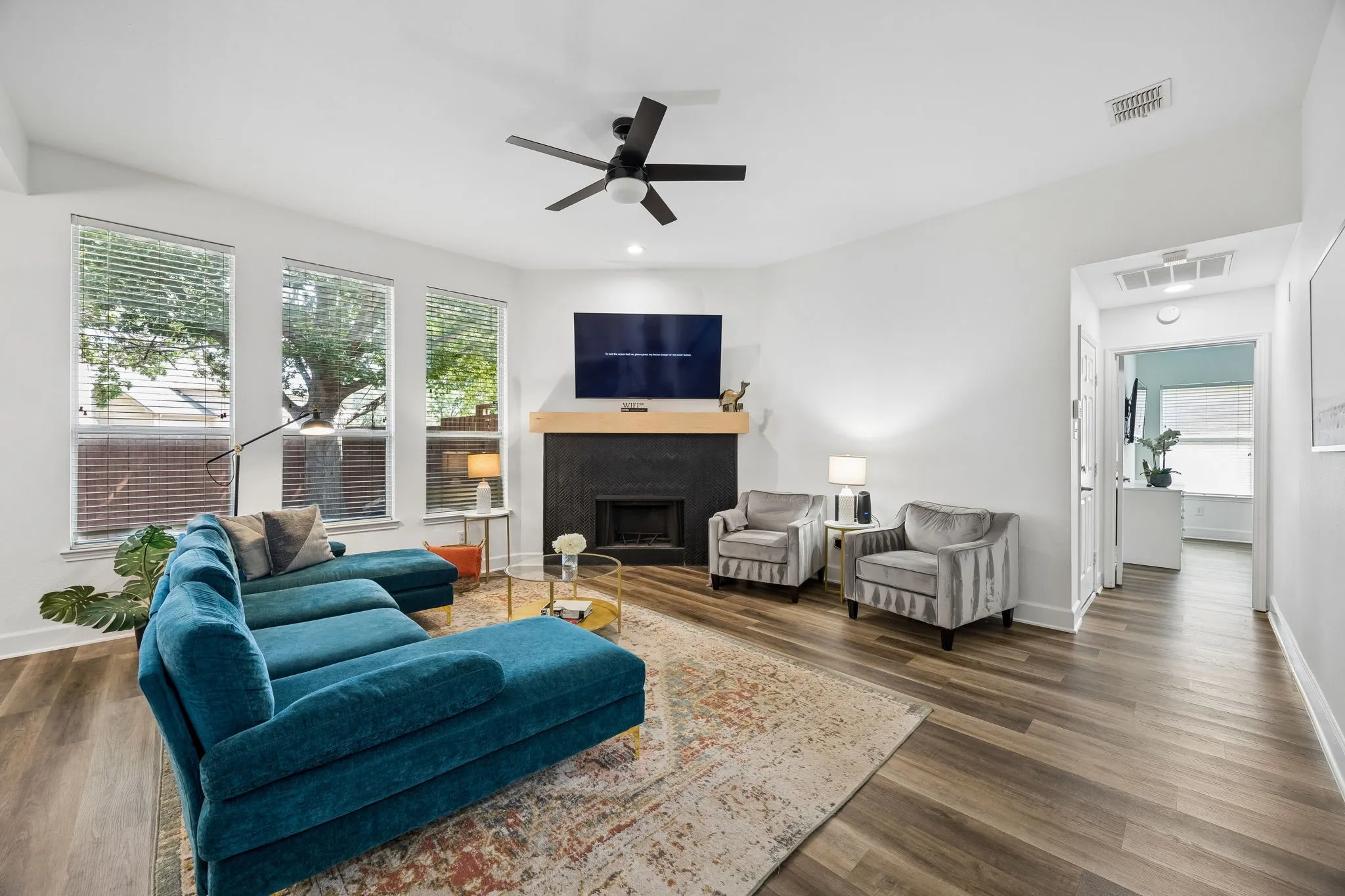 Vaulted ceilings with a ceiling fan, wood-finish flooring, multiple large windows with blinds, black-painted fireplace with a wood mantle, and white walls