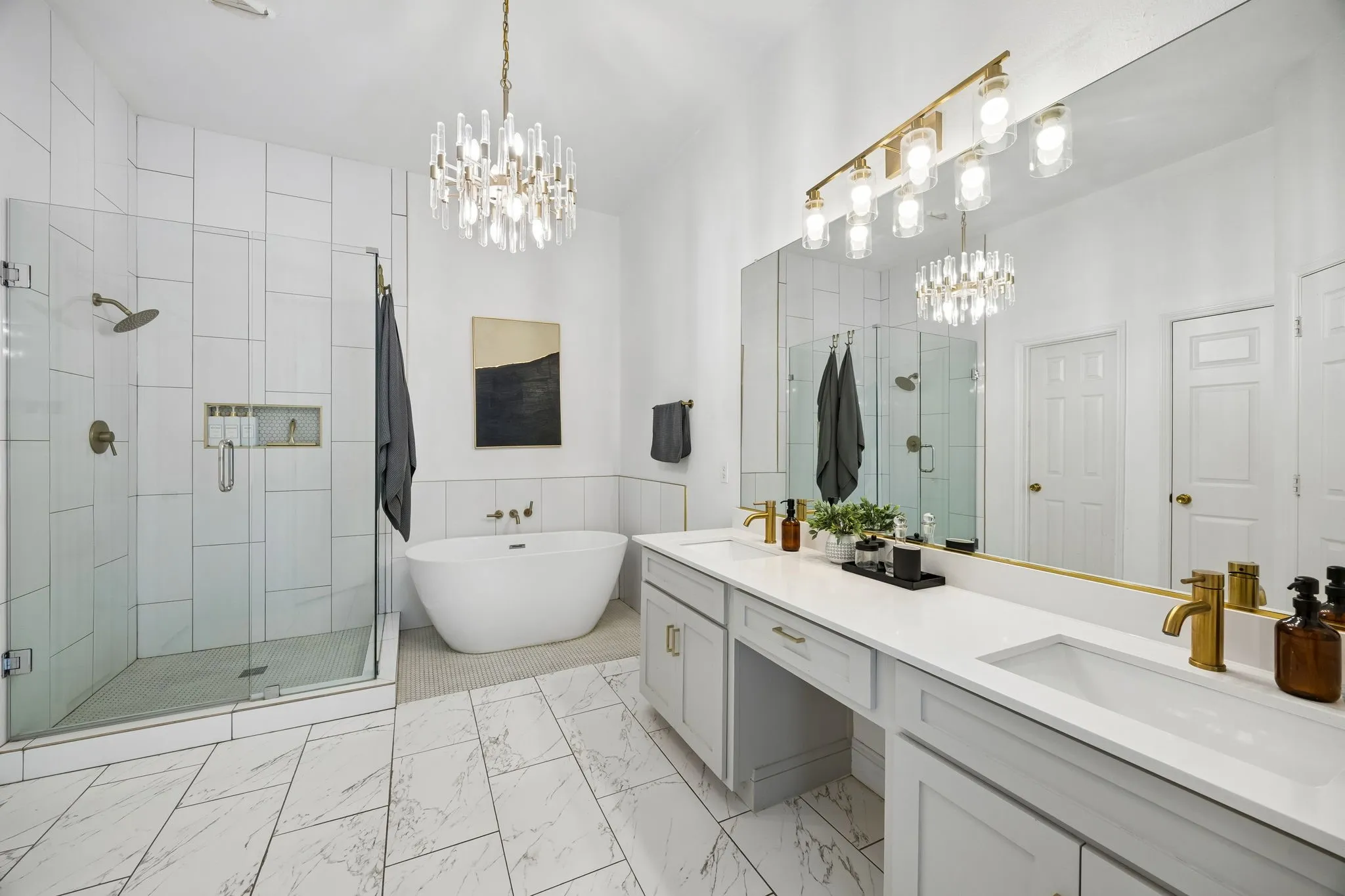 Freestanding soaking tub, frameless glass shower enclosure with tile surround, dual vanity with undermount sinks, and a crystal chandelier