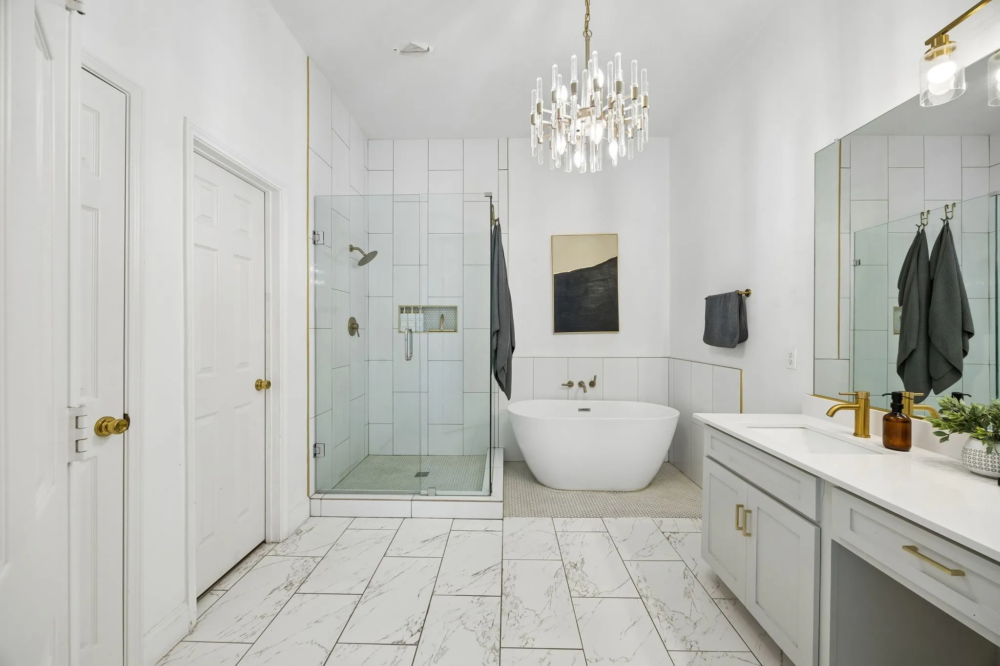 Freestanding soaking tub, glass-enclosed shower, dual vanity with undermount sink, marble-look tile flooring, and a crystal chandelier