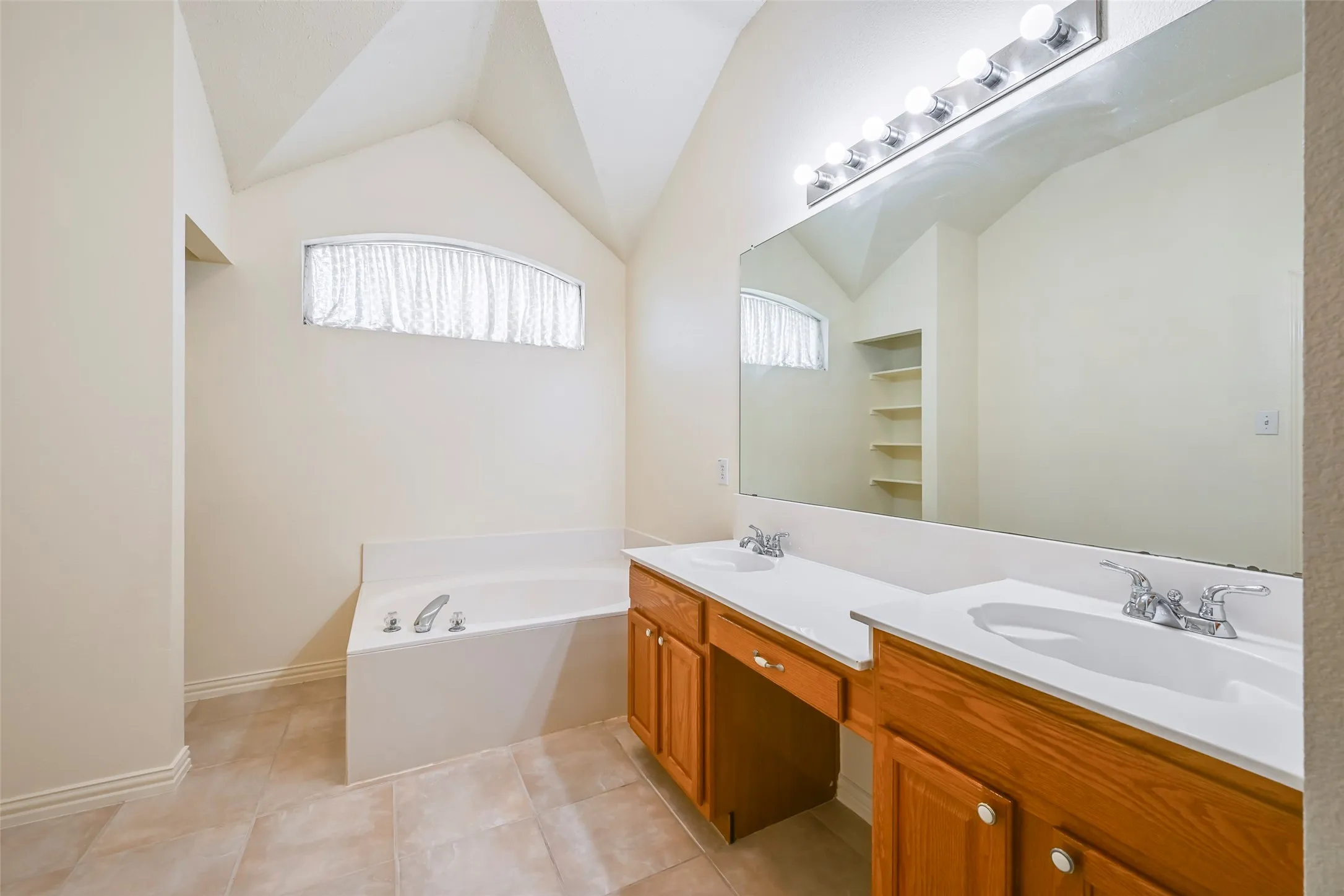 Dual vanity with wood cabinetry, recessed bathtub, arched window, and vaulted ceiling