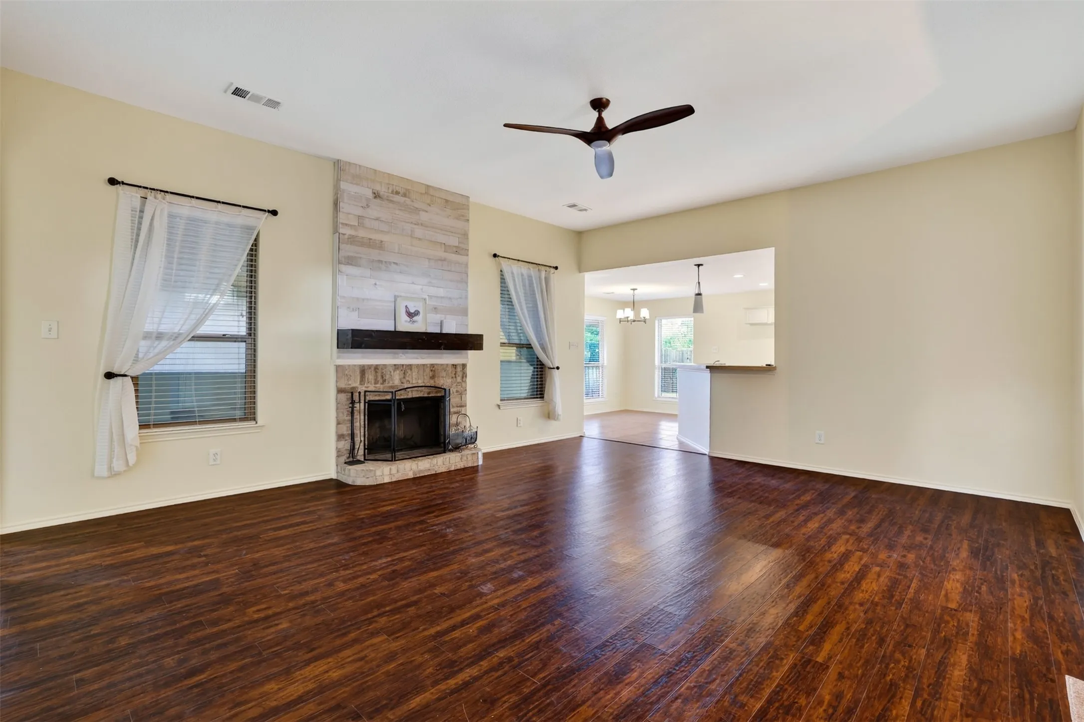 Spacious living area featuring wood-finish flooring, a brick fireplace with a wood mantel, and a ceiling fan all open to a lovely kitchen