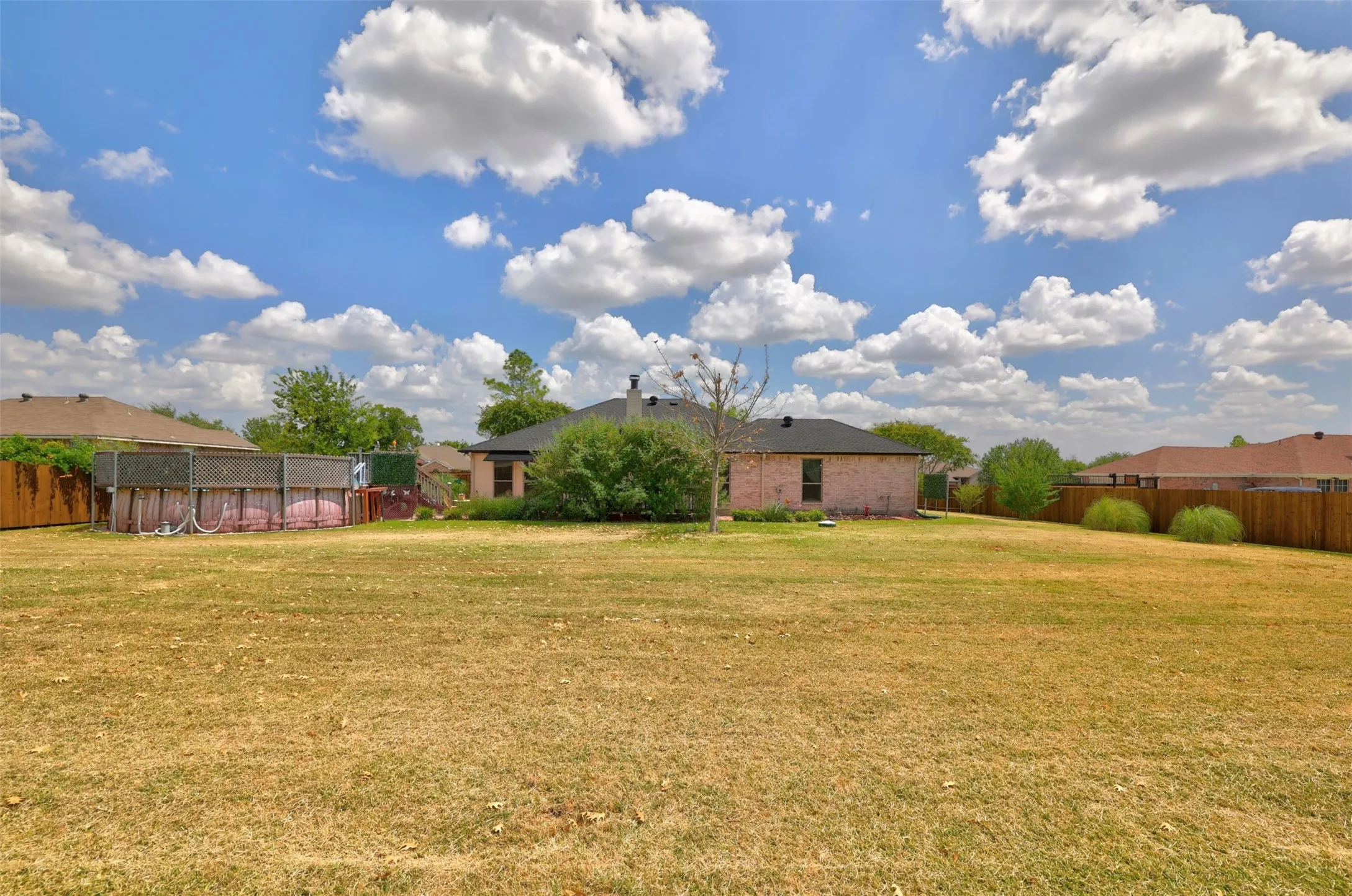 Expansive grassy outdoor space featuring a wood fence, mature trees, a brick exterior and pool