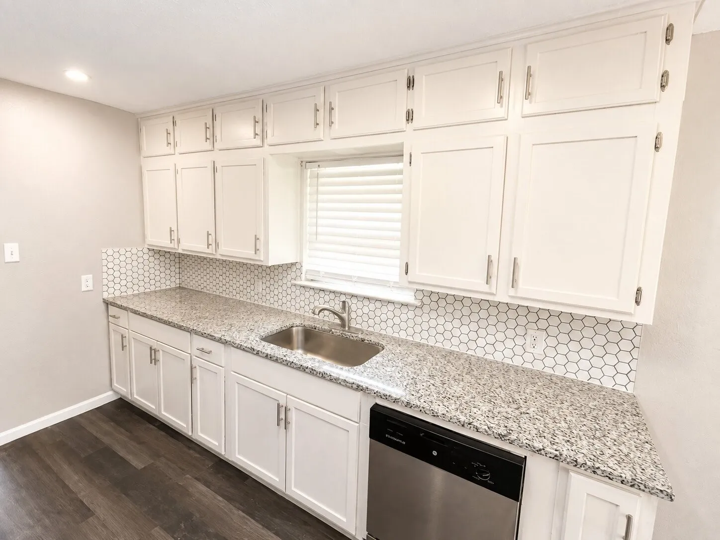 White shaker-style cabinetry with brushed nickel hardware, light-toned speckled countertops, hexagonal tile backsplash, stainless steel sink, and wood-finish flooring