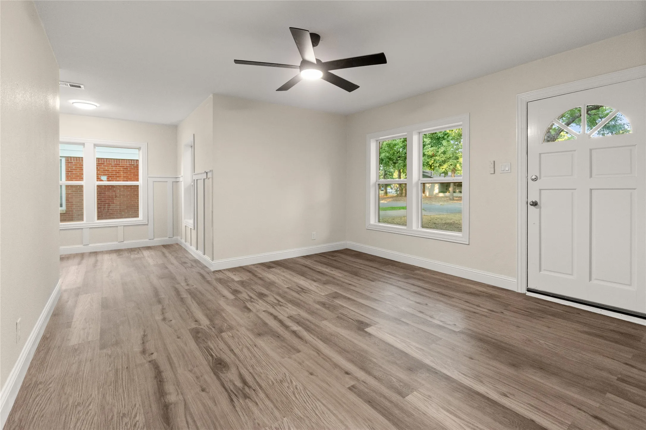 Living room with wood-finish flooring, white paneled interior door with arched glass detail, dual windows, modern ceiling fan with integrated lighting, and neutral wall tones