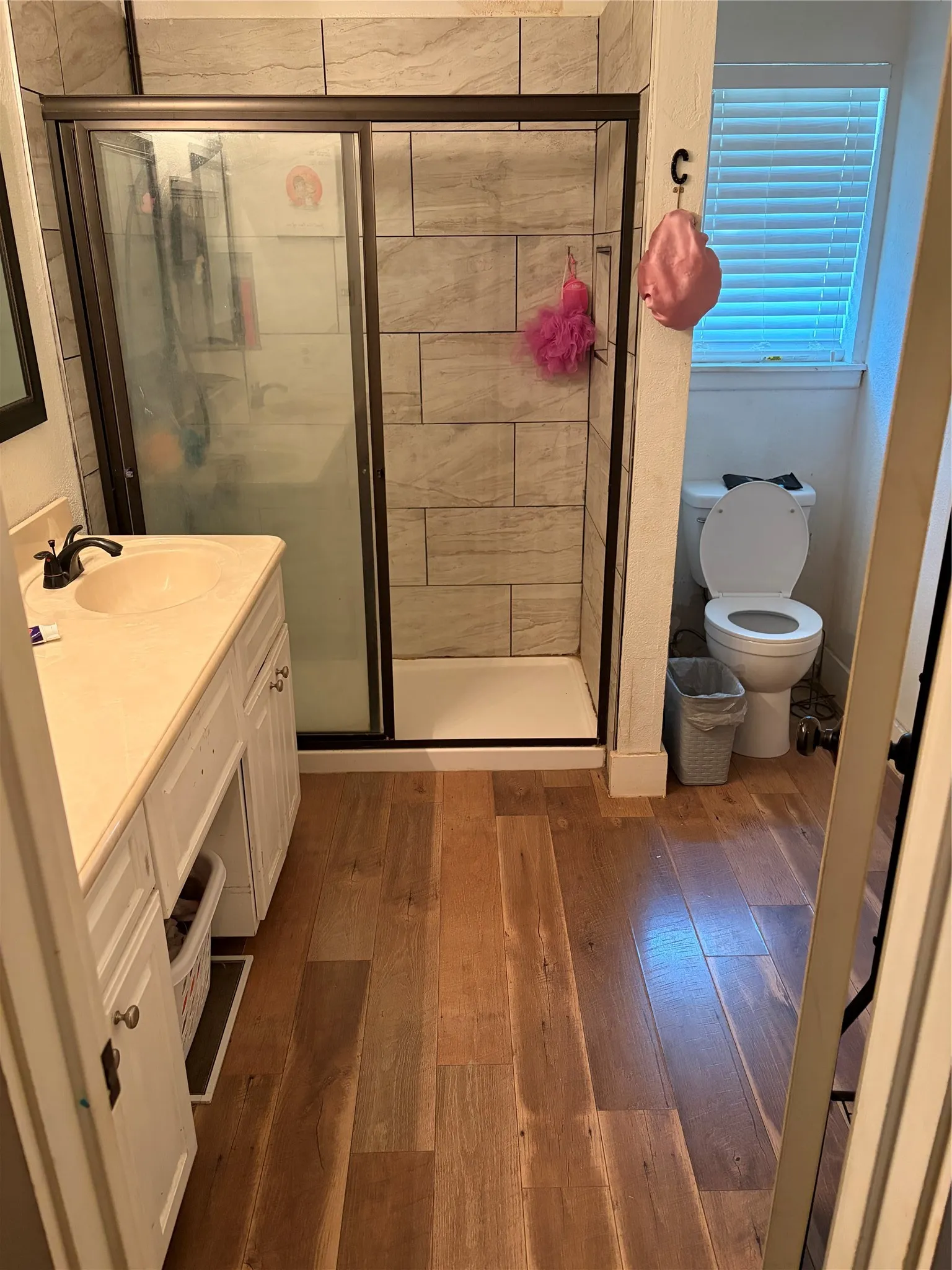 Wood-finish flooring, white vanity with integrated sink, tiled shower surround, sliding glass shower door, and privacy window blinds