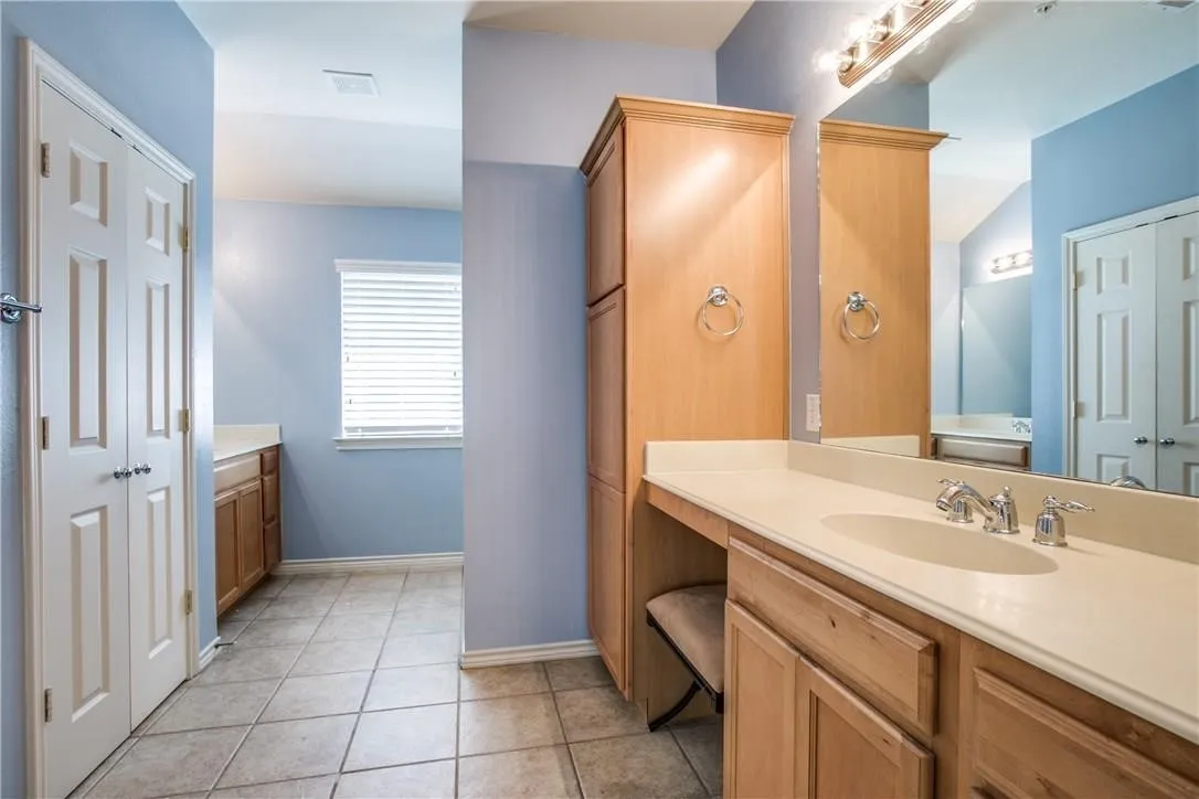 Light-toned wood-finish cabinetry, tile flooring, vanity mirror, and window with horizontal blinds