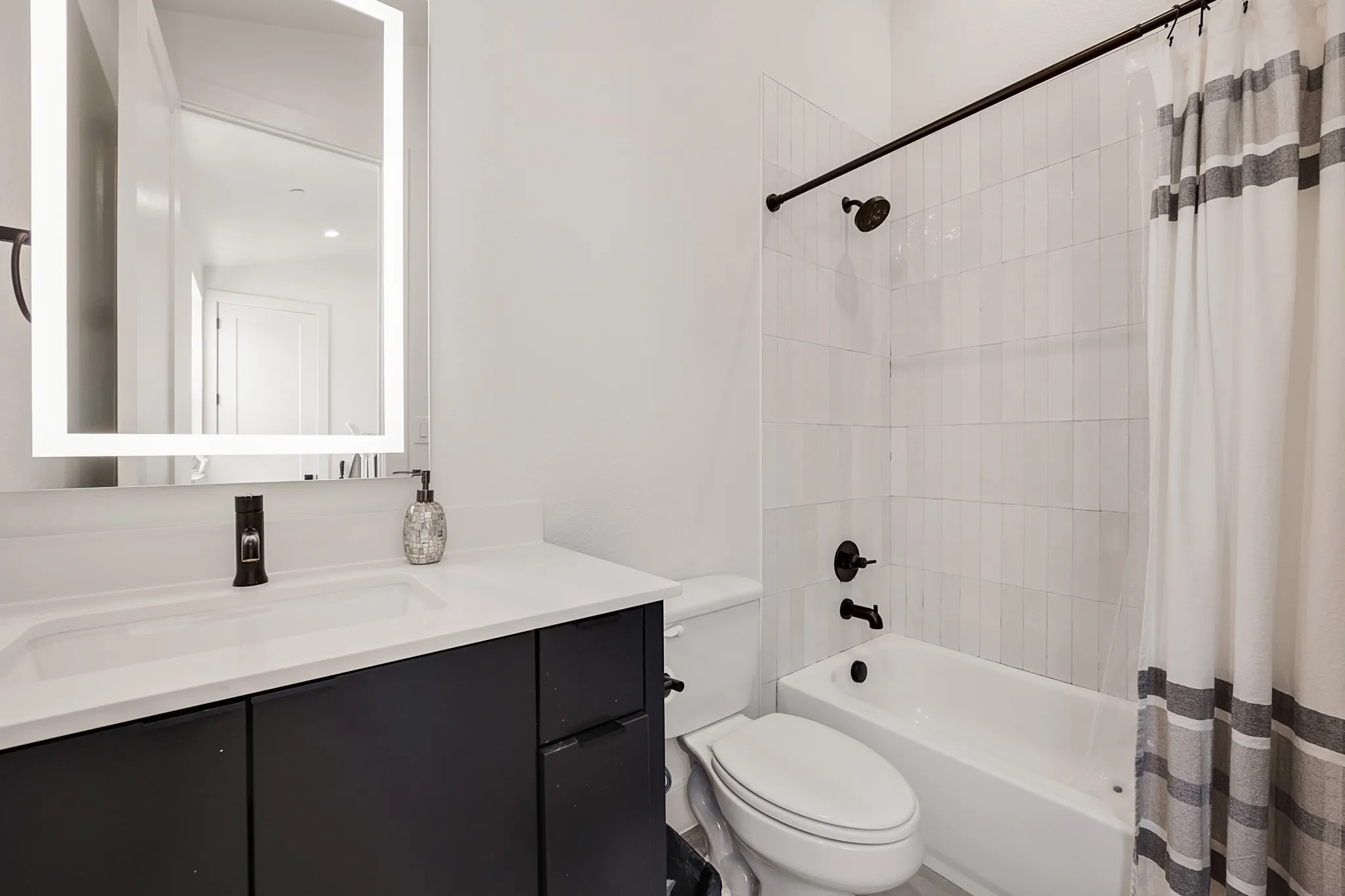 Black vanity with integrated sink, white countertop, illuminated mirror, and a white tile shower surround with black fixtures