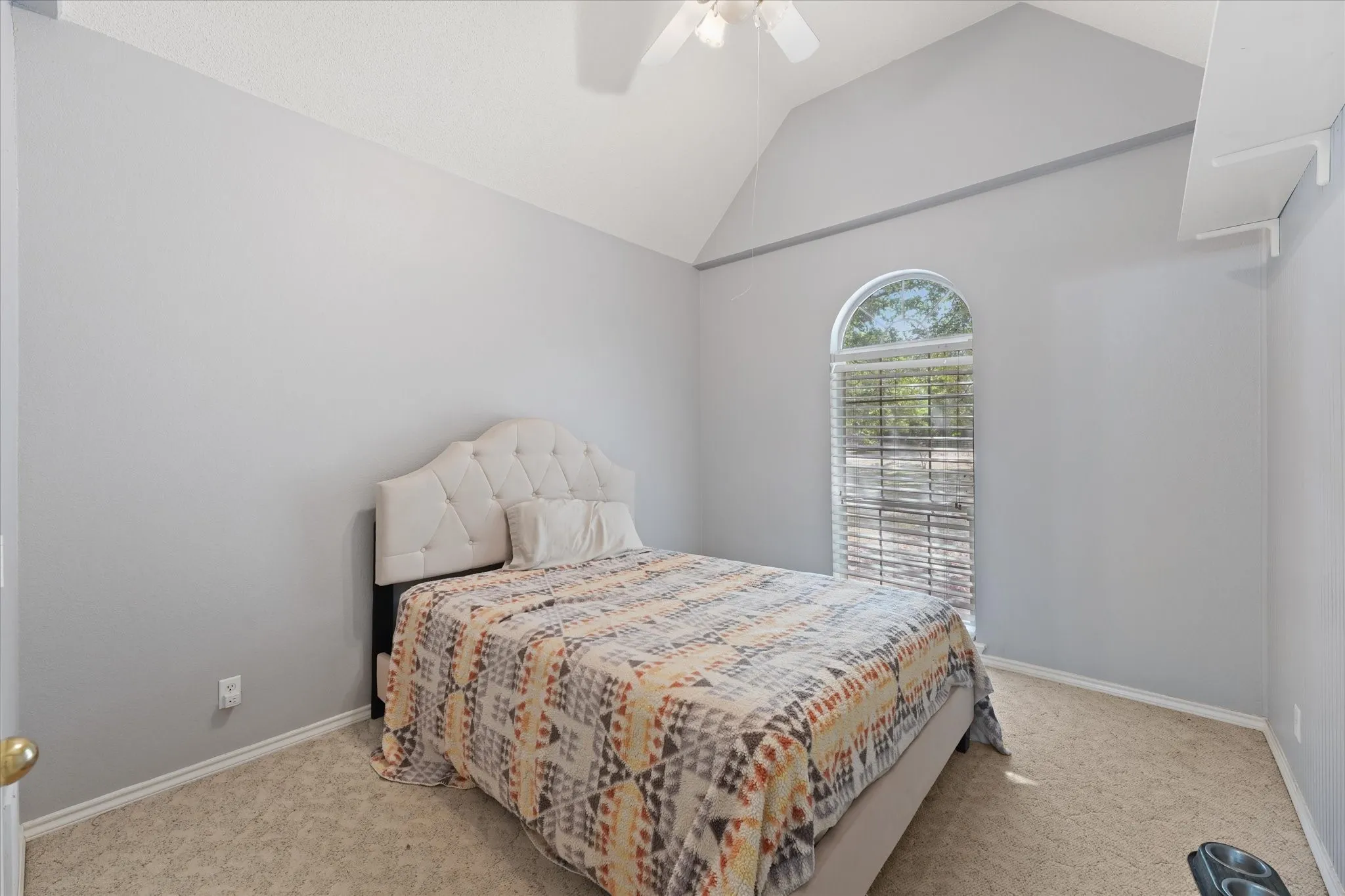 Bright secondary bedroom with vaulted ceiling, ceiling fan, neutral carpet flooring, and an arched window with plantation shutters providing natural light and architectural interest