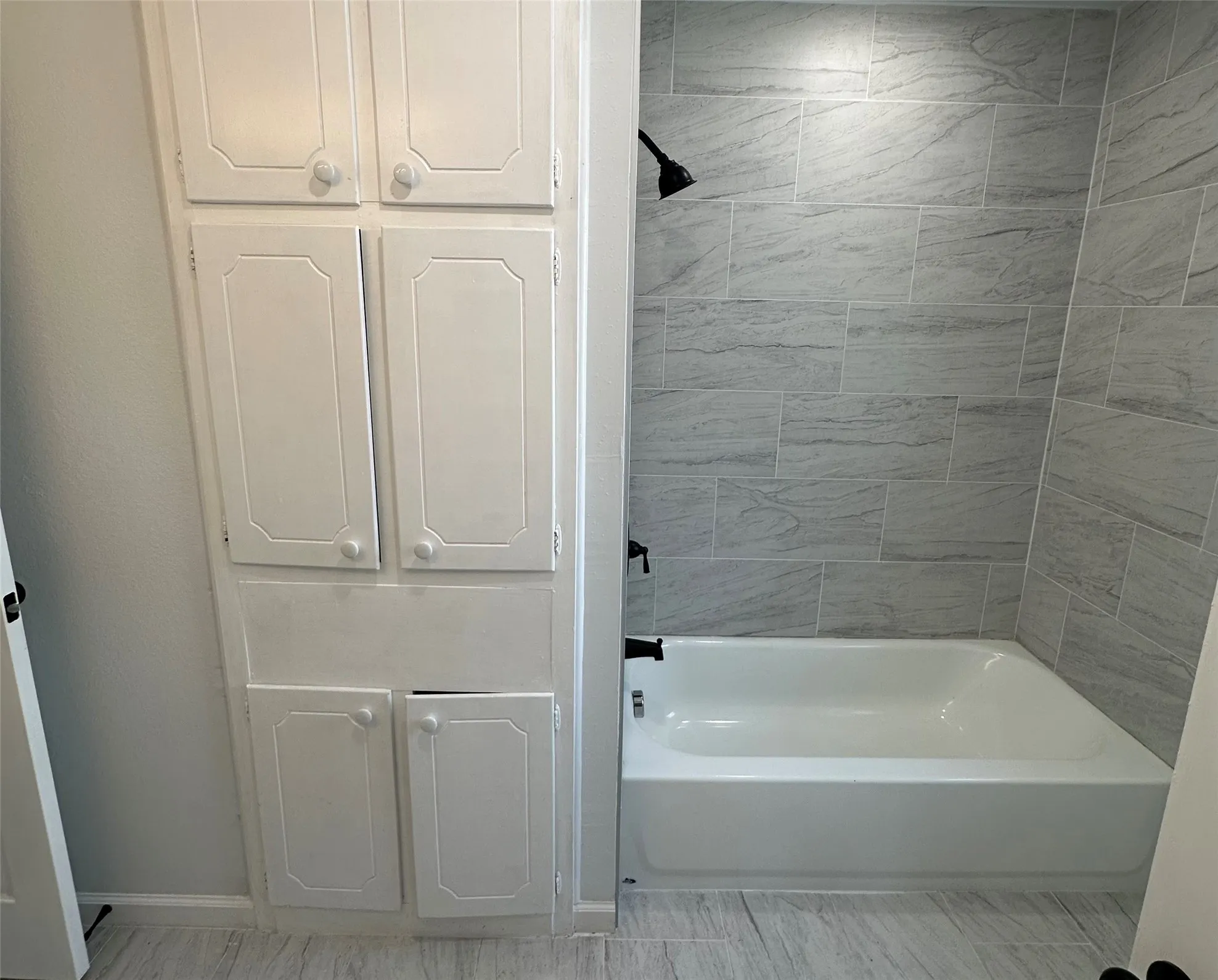 Built-in storage cabinetry, white soaking tub, light gray tile flooring, matte black shower fixtures, light gray shower wall tiling