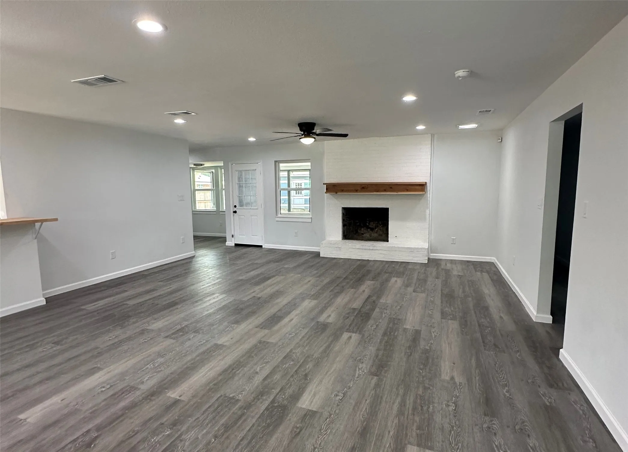 Spacious living area featuring wood-finish flooring, recessed lighting, and a white-painted brick fireplace with a wood mantel