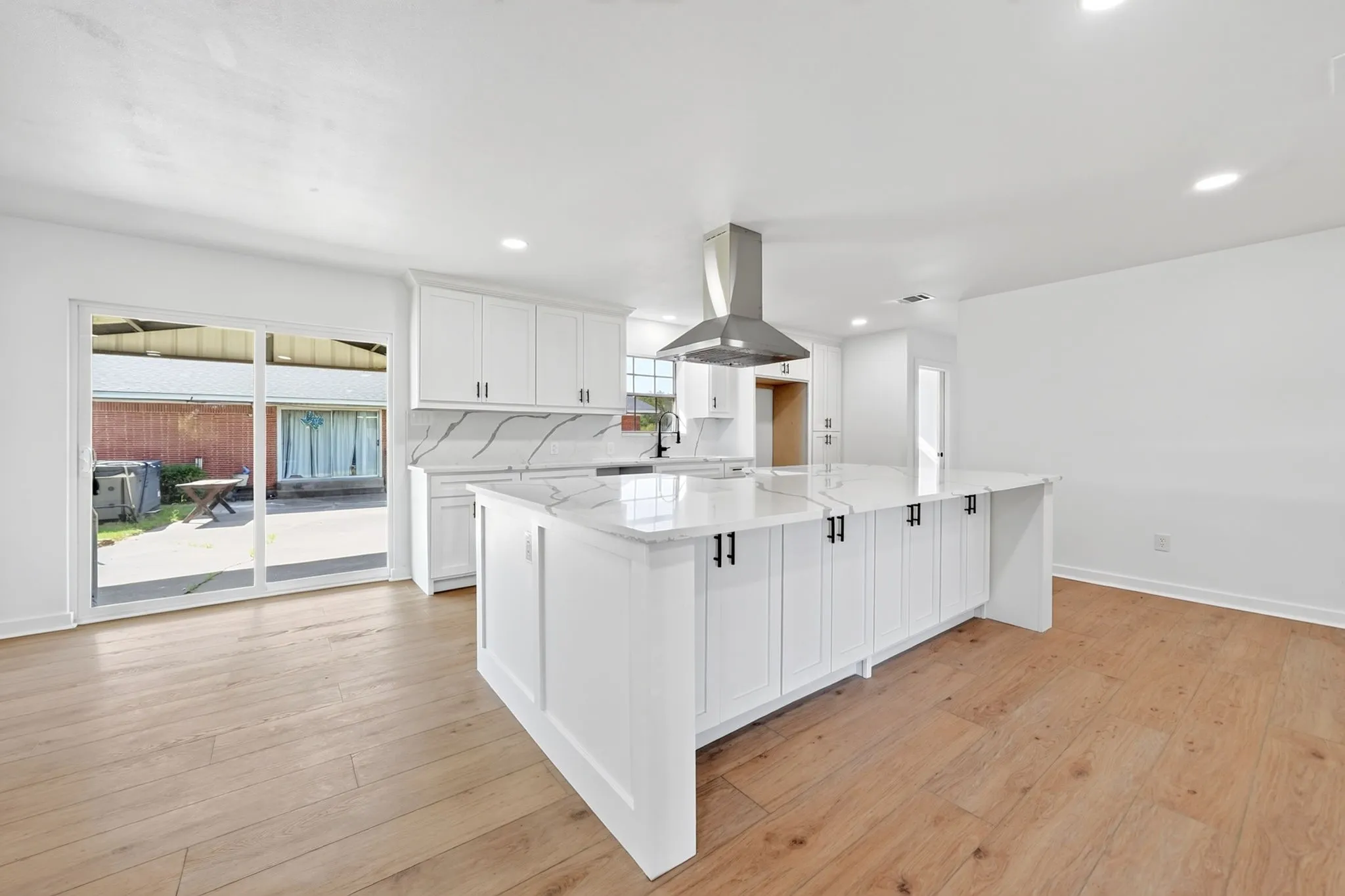Spacious kitchen with a large central island, white cabinetry, integrated range hood, wood-finish flooring, and sliding glass doors