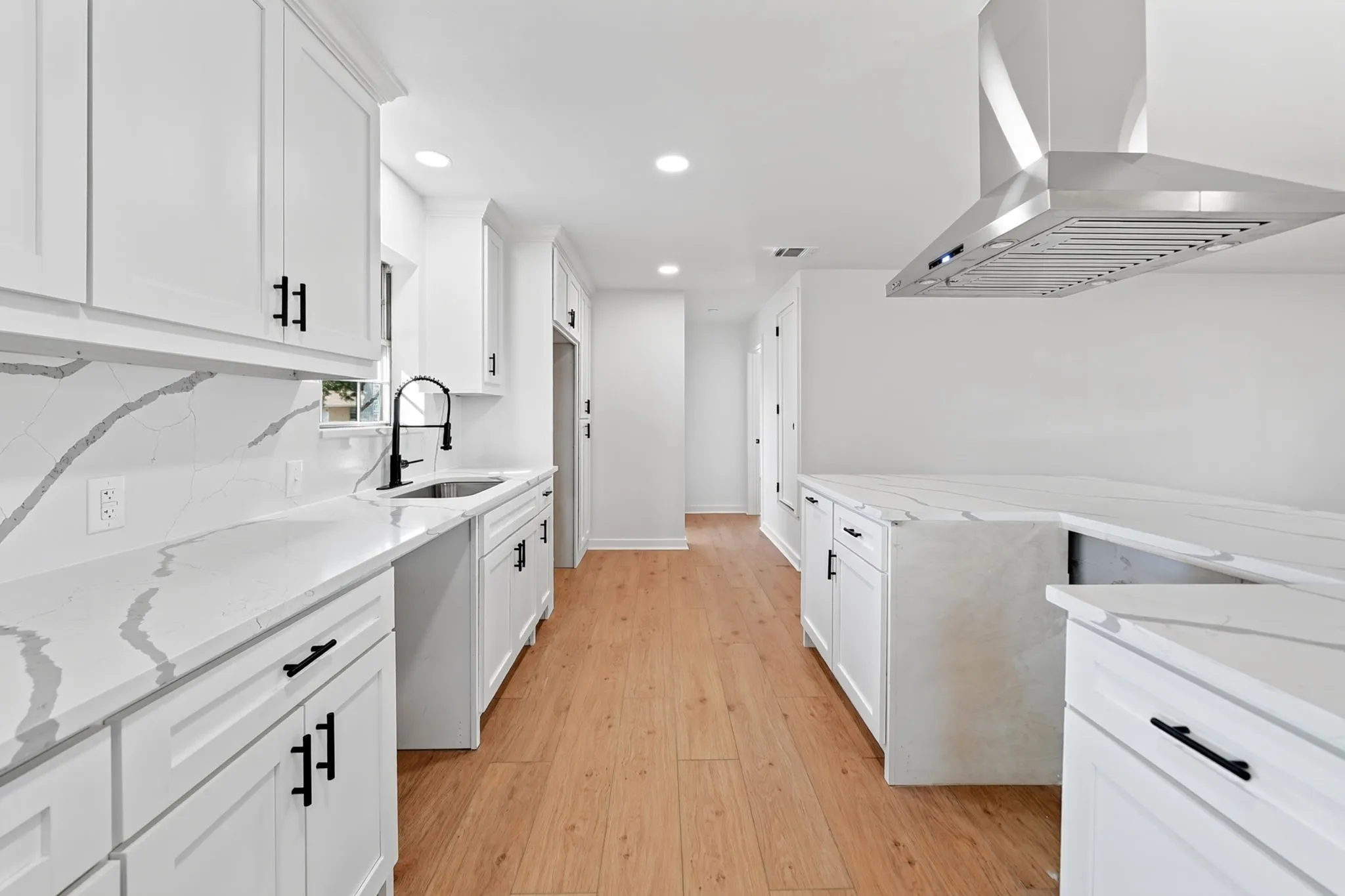 Galley kitchen featuring white cabinetry with black hardware, quartz countertops with subtle veining, stainless steel appliances including a professional-style range hood, undermount sink with contemporary faucet, recessed lighting, and warm wood-finish flooring throughout