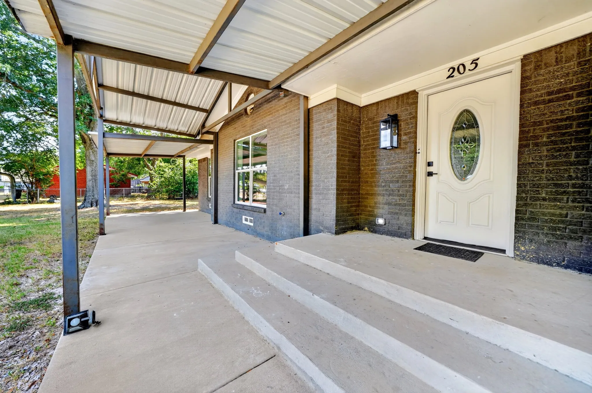 Covered concrete patio with metal roof structure, brick exterior, and decorative entry door with oval glass insert