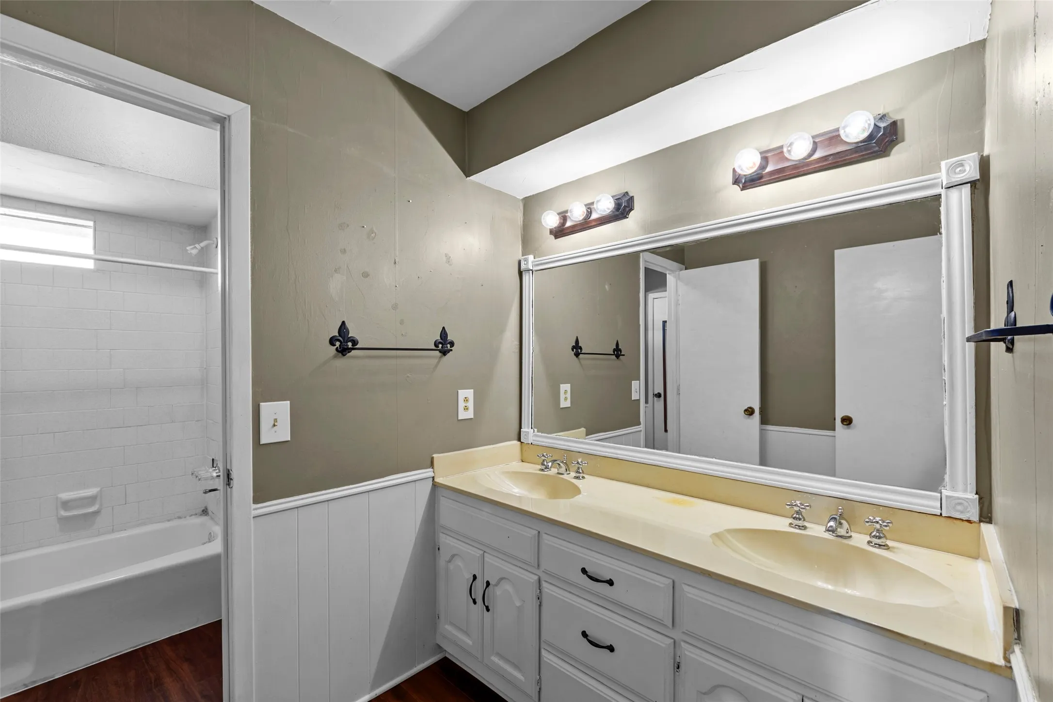 Dual vanity with integrated sinks and white cabinetry, large framed mirror, wood-finish flooring, and a tiled tub shower