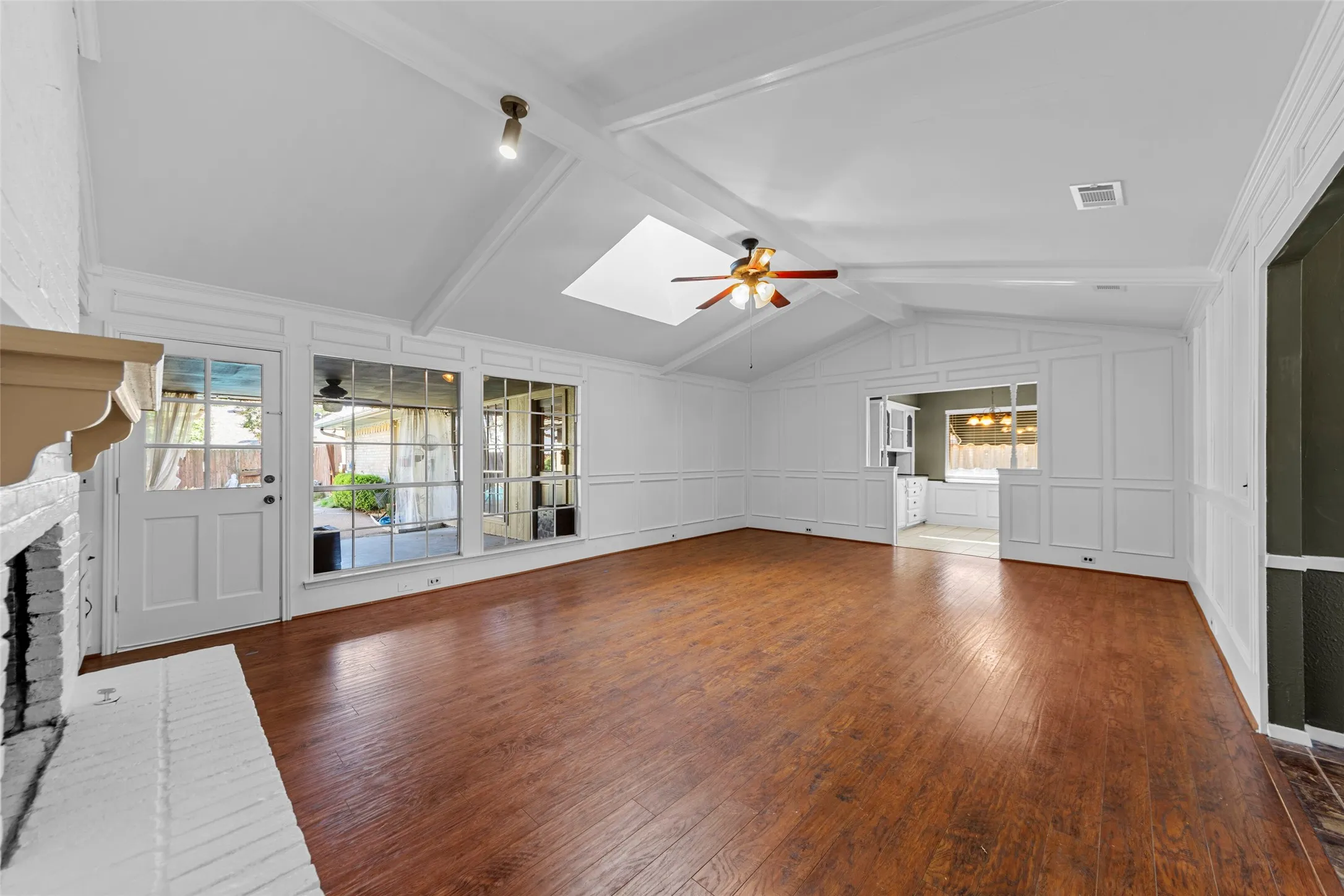 Vaulted ceiling with exposed beams, wood-finish flooring, white painted brick fireplace, paneled accent walls, and a ceiling fan with integrated lighting