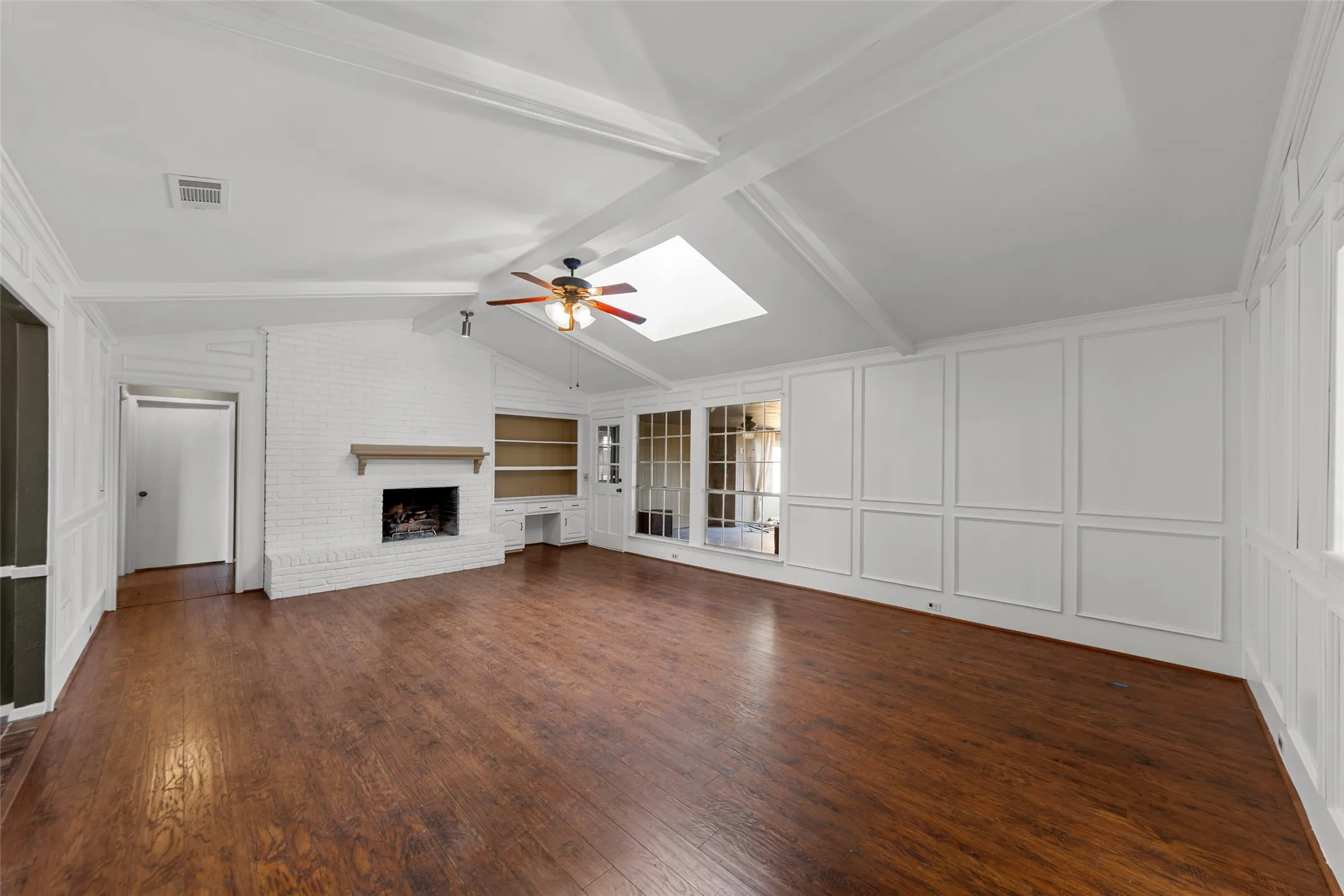 Vaulted ceilings with exposed beams, wood-finish flooring, white brick fireplace with mantel, built-in shelving, and skylight