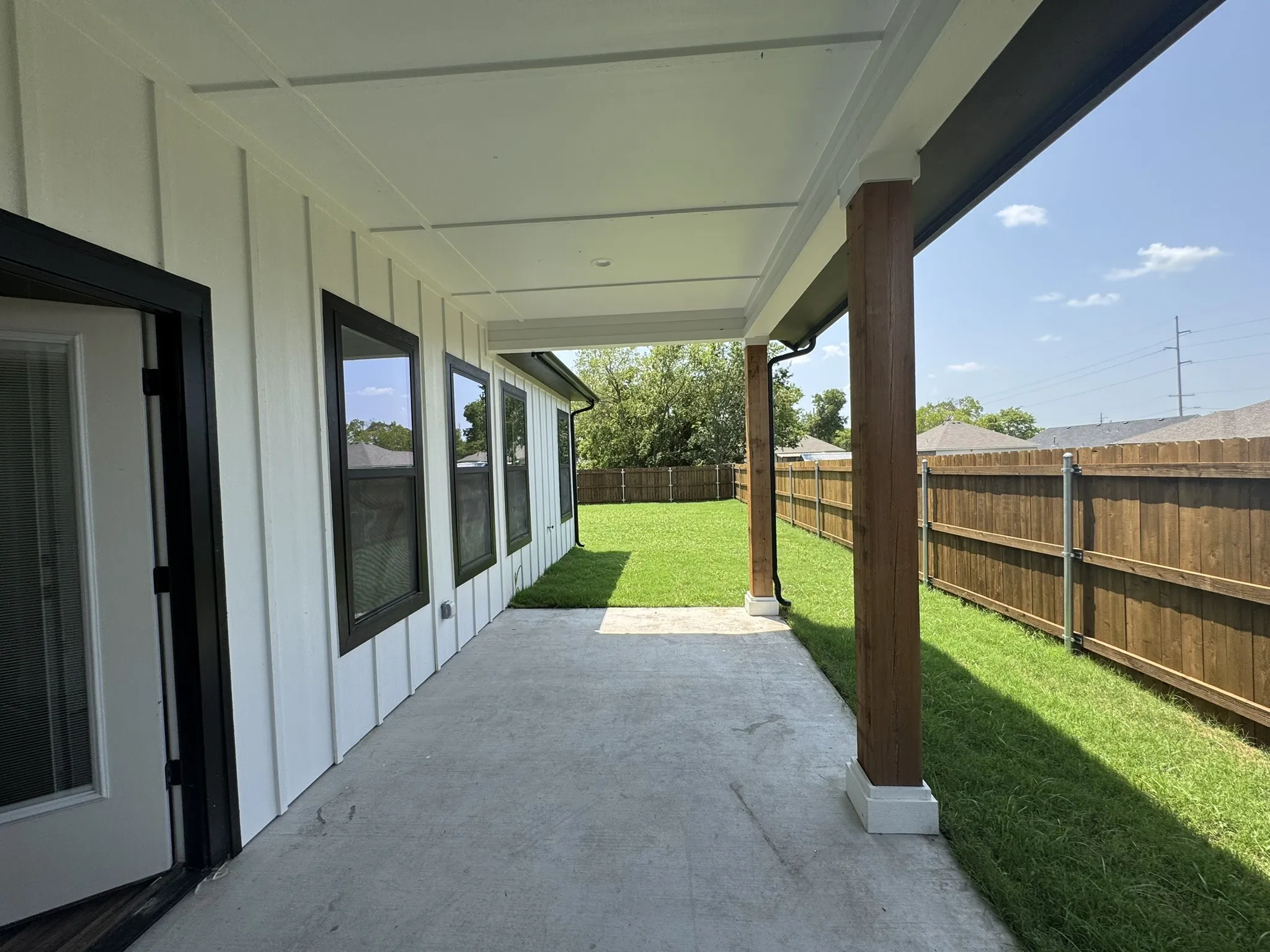 Covered patio featuring a concrete surface, wood-finish support columns, white board and batten siding, dark-framed windows, and a wood privacy fence