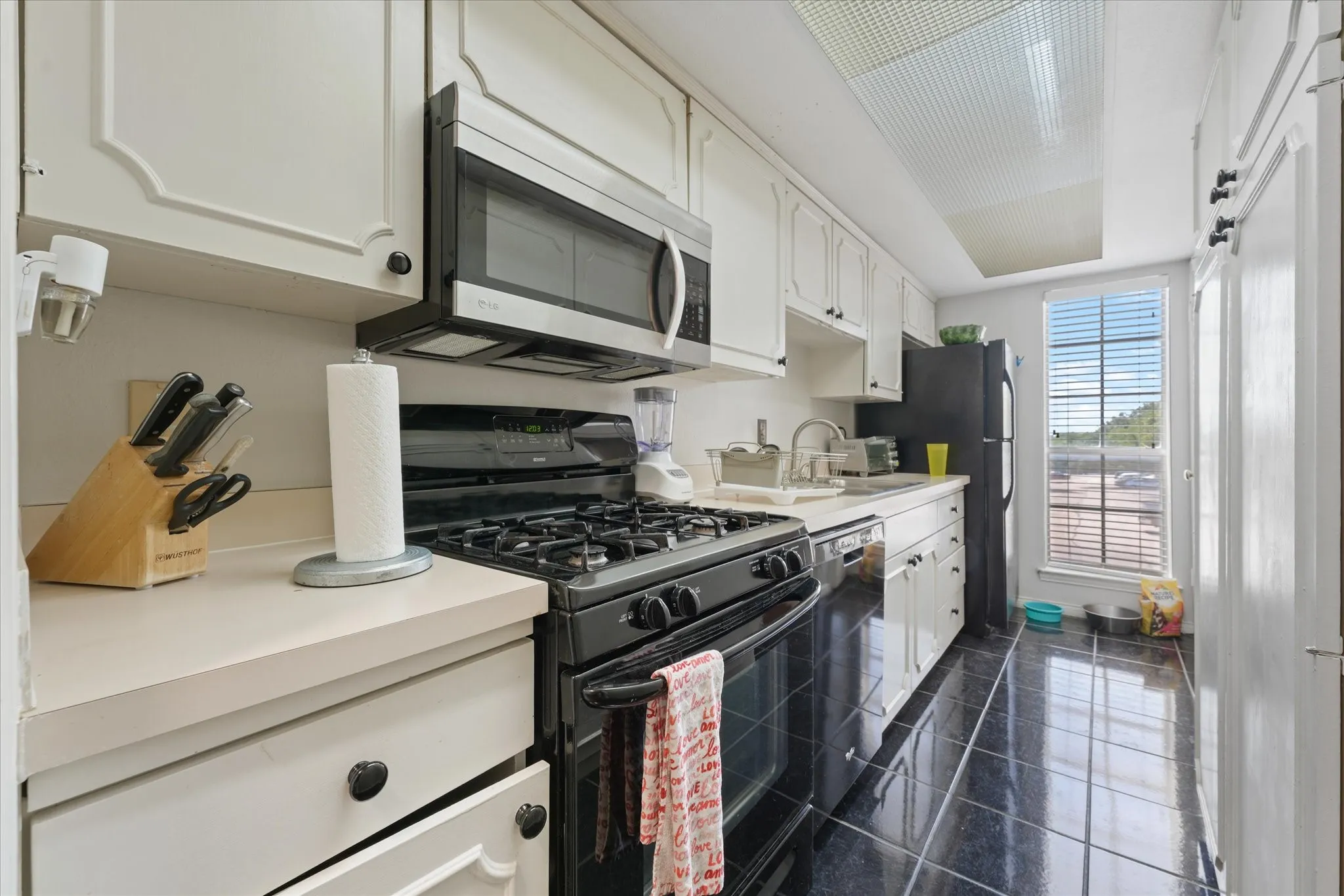 Galley kitchen featuring black tile flooring, white cabinetry with black hardware, white countertops, stainless steel microwave, and a gas range
