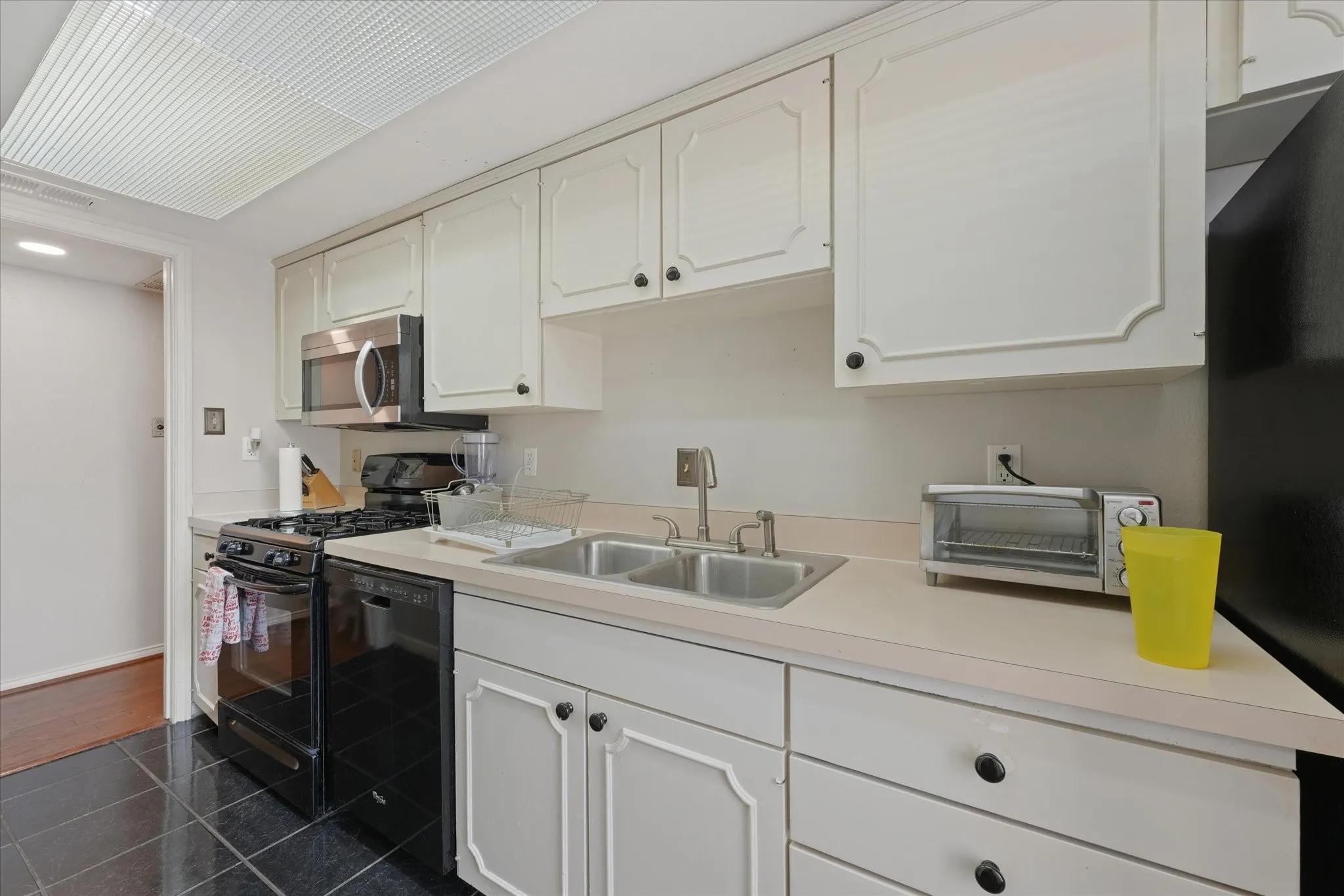 Galley kitchen featuring white cabinetry, a double basin stainless steel sink, black tile flooring, and a stainless steel microwave
