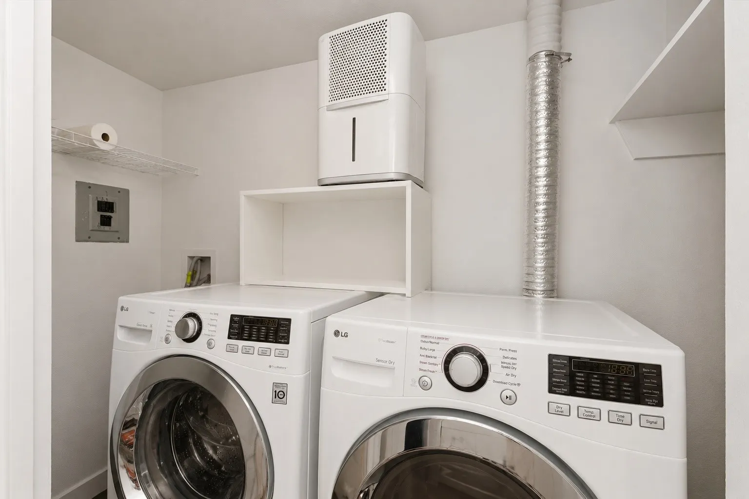 White laundry appliances with front-loading design, wall-mounted shelving, and overhead ductwork