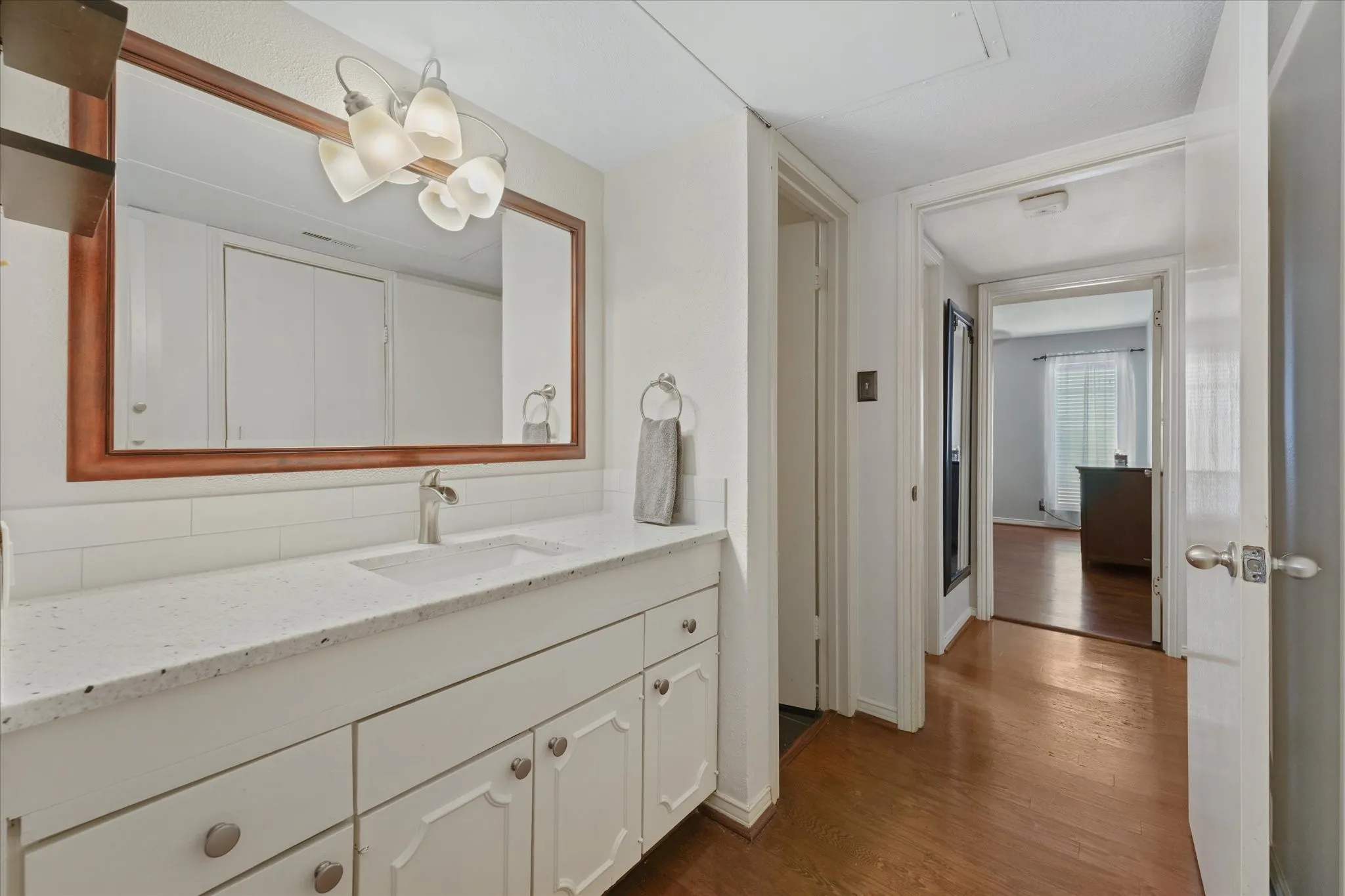 White vanity with integrated sink and chrome faucet, large framed mirror, vanity light fixture, white subway tile backsplash, and wood-finish flooring