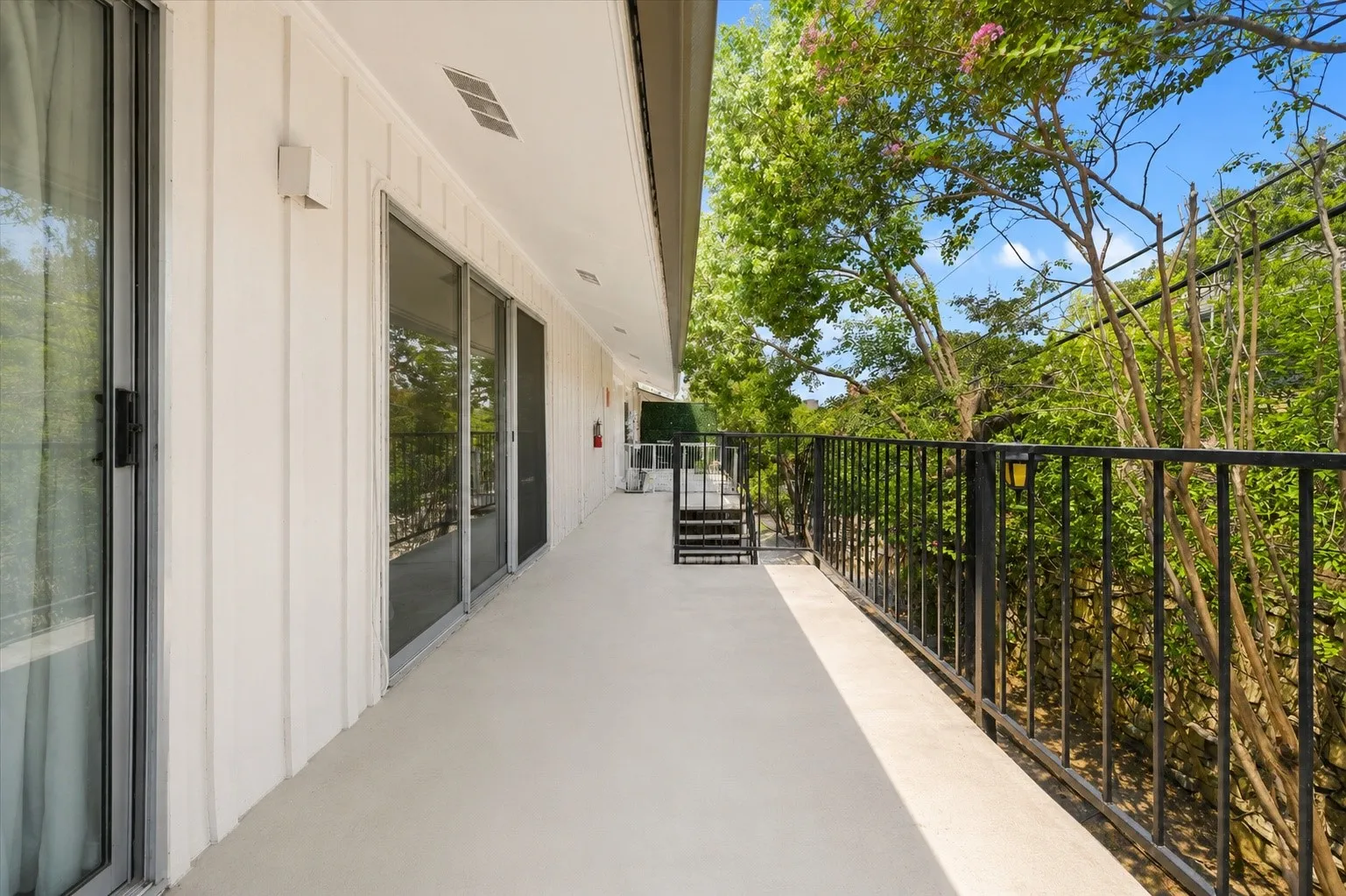 Expansive outdoor balcony with a light-toned surface, black metal railing, and multiple sliding glass doors