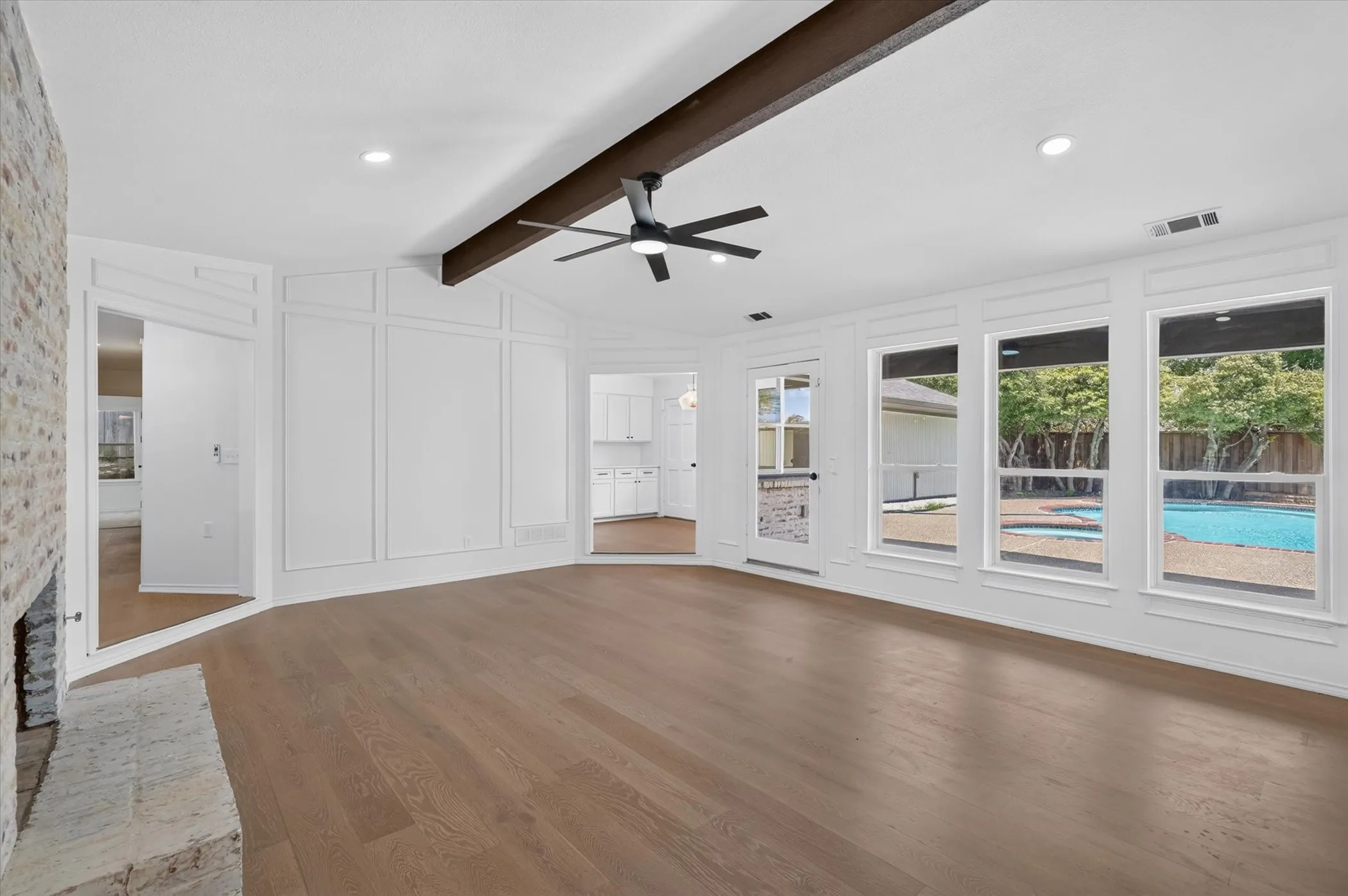 Vaulted ceiling with exposed wood beam, wood-finish flooring, stone fireplace, decorative paneling, and multiple windows overlooking a swimming pool