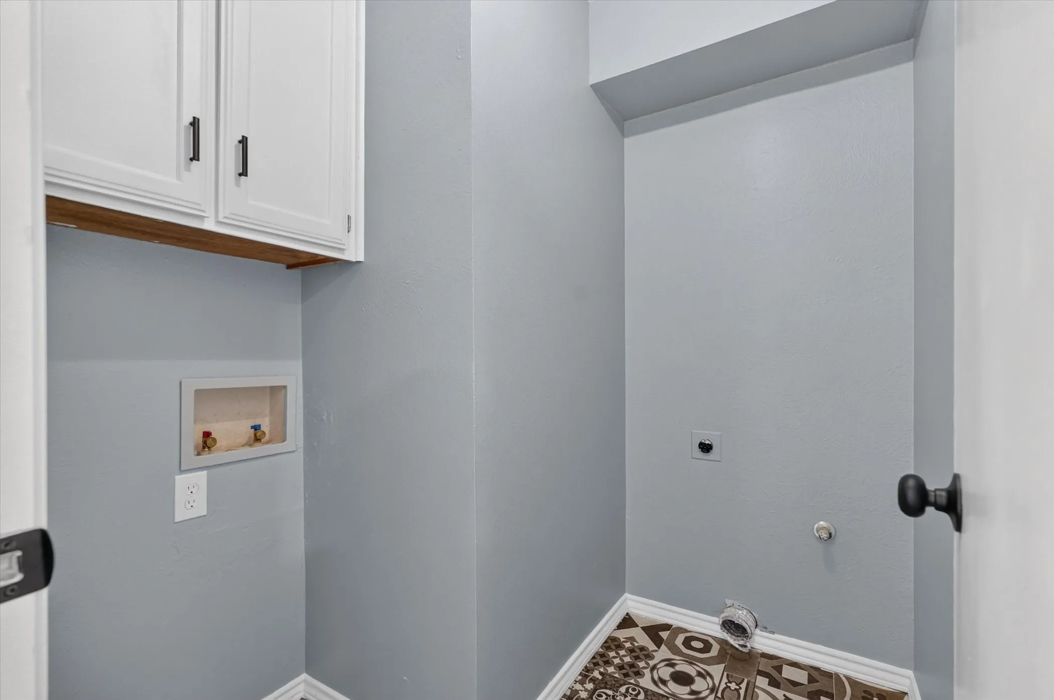 White shaker cabinetry with dark hardware, wood-finish shelving, utility hookups, and patterned tile flooring