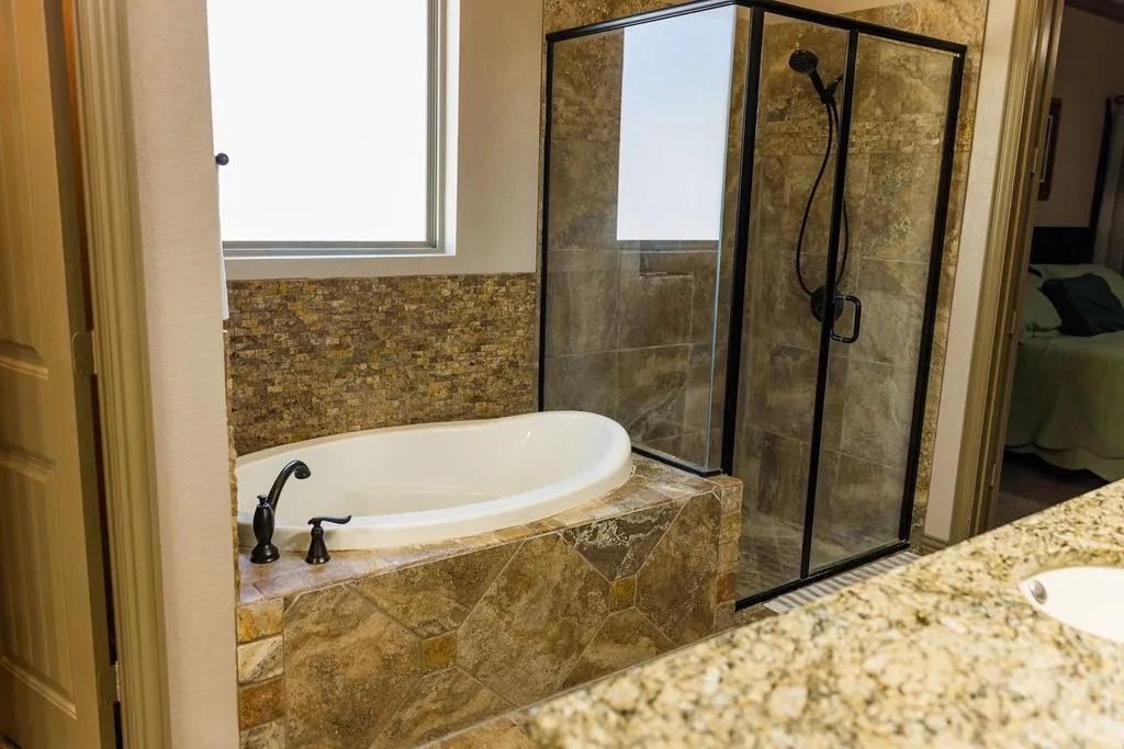 Soaking tub with bronze fixtures, glass-enclosed shower, stone tile surround, and a window for natural light